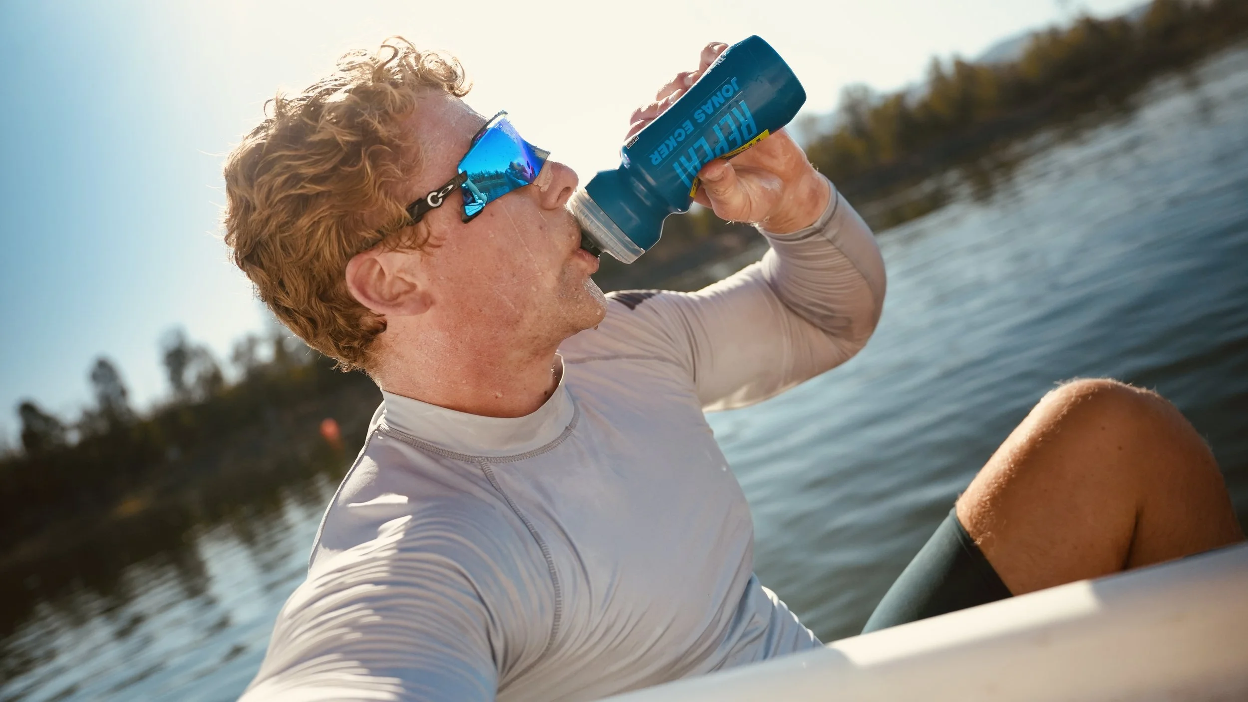 A person with curly red hair and sunglasses drinks from a fish bait can while sitting in a boat on the water.