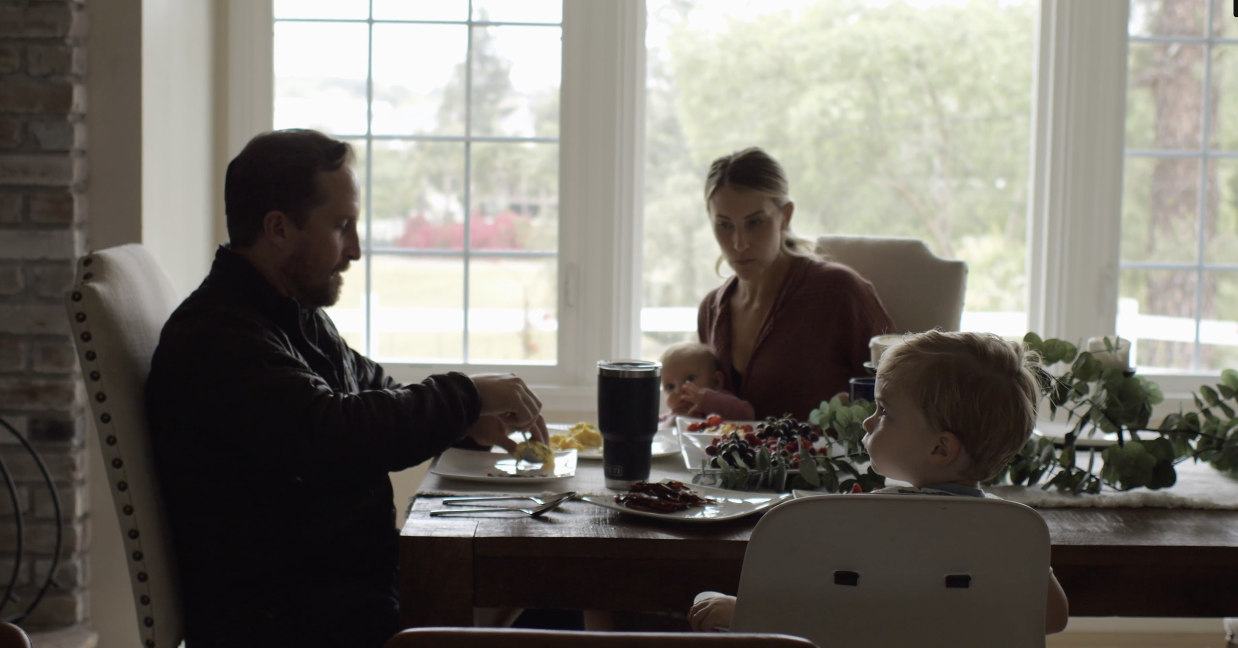 Family having breakfast at a dining table in a bright kitchen with large windows and greenery outside.