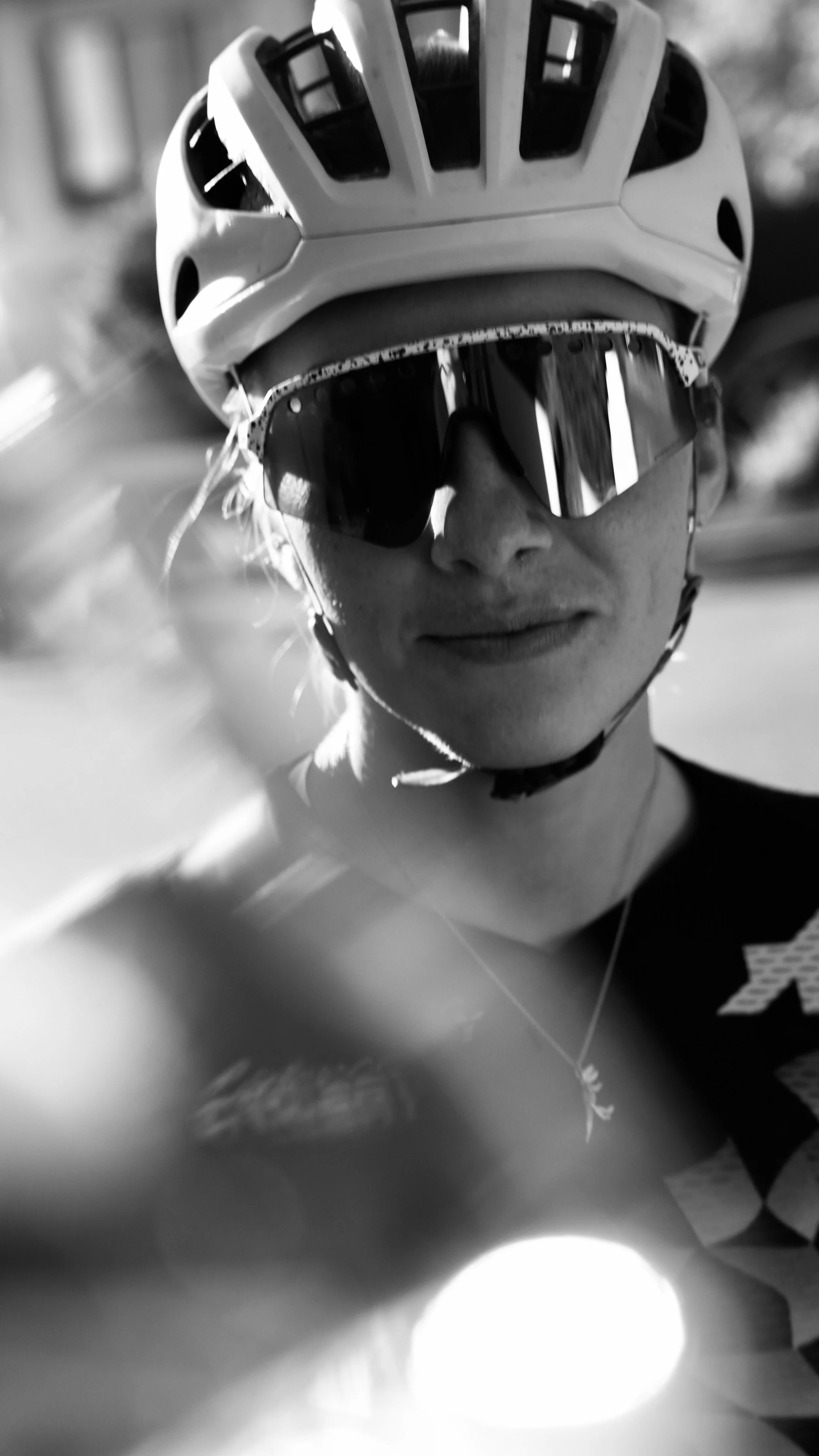 Close-up black and white photo of a smiling woman wearing a bike helmet and sunglasses, with light reflecting off her sunglasses.