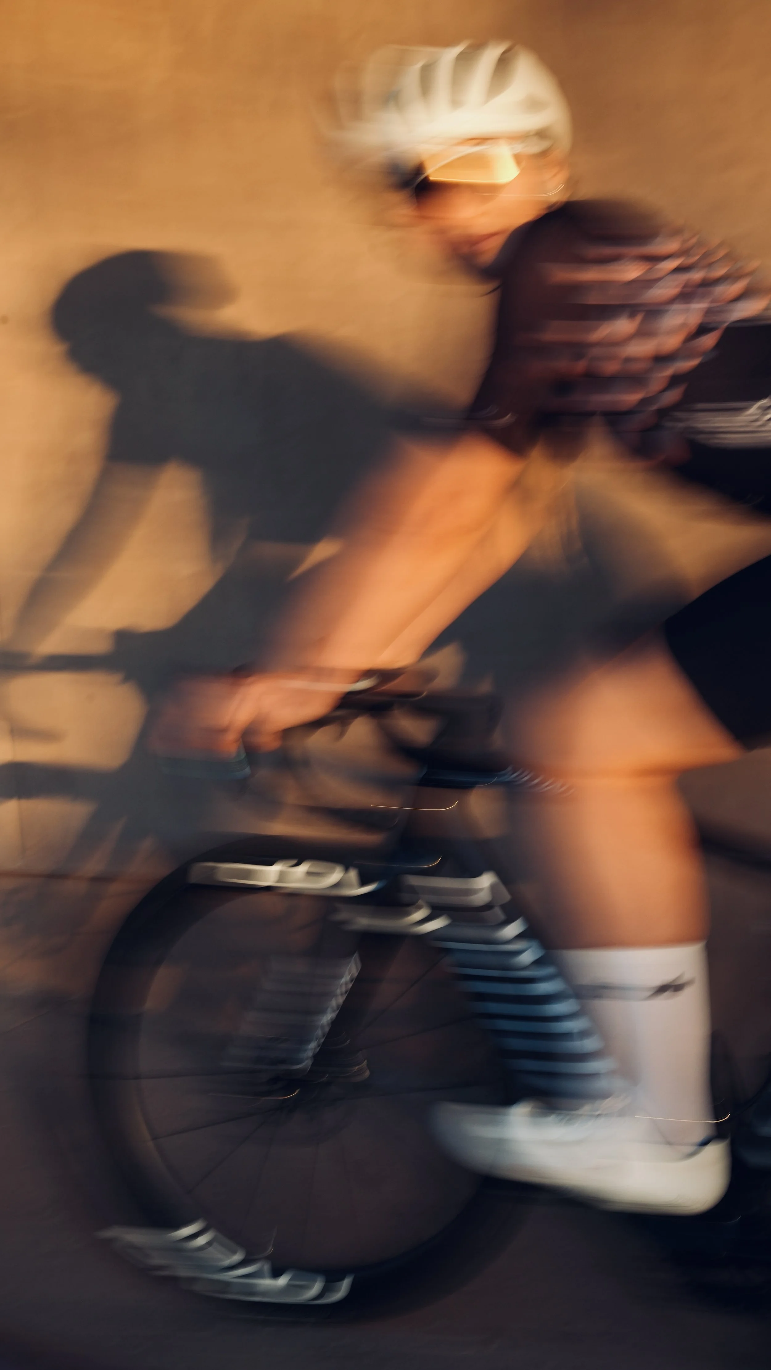 Blurred photo of a cyclist wearing a white helmet and striped socks, riding a bike indoors, with a brown wall in the background.