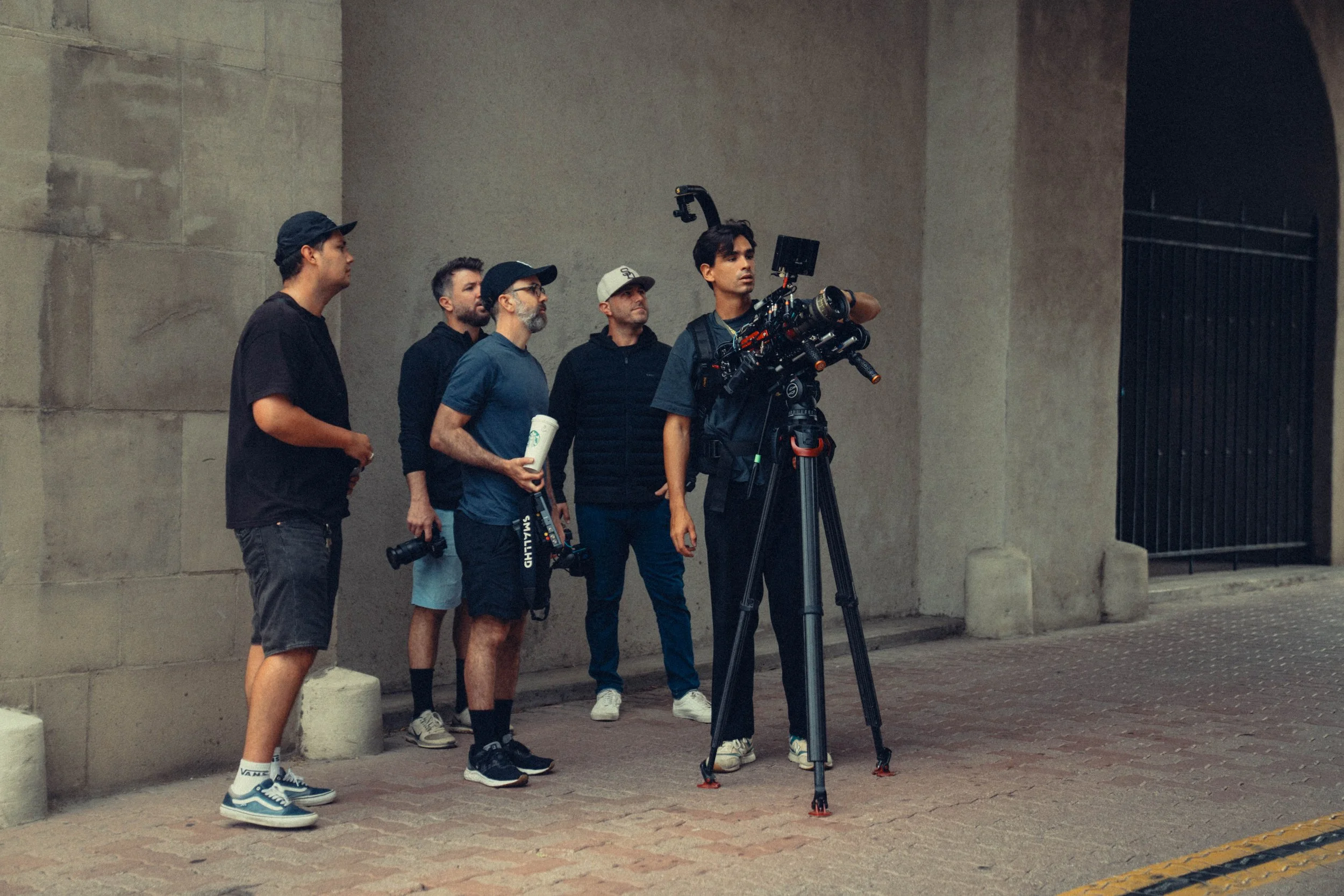 Group of five men operating professional film or video camera equipment on city sidewalk at night.