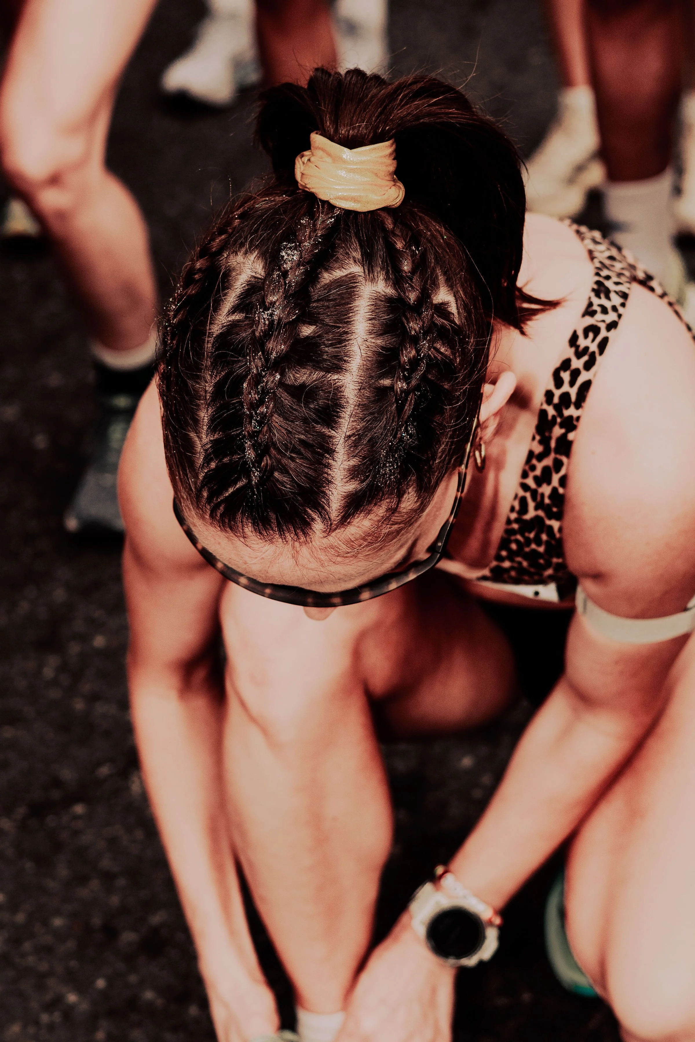 A person with dark hair in braids wearing a beige scrunchie, crouching with others around on a dark surface, wearing athletic gear.