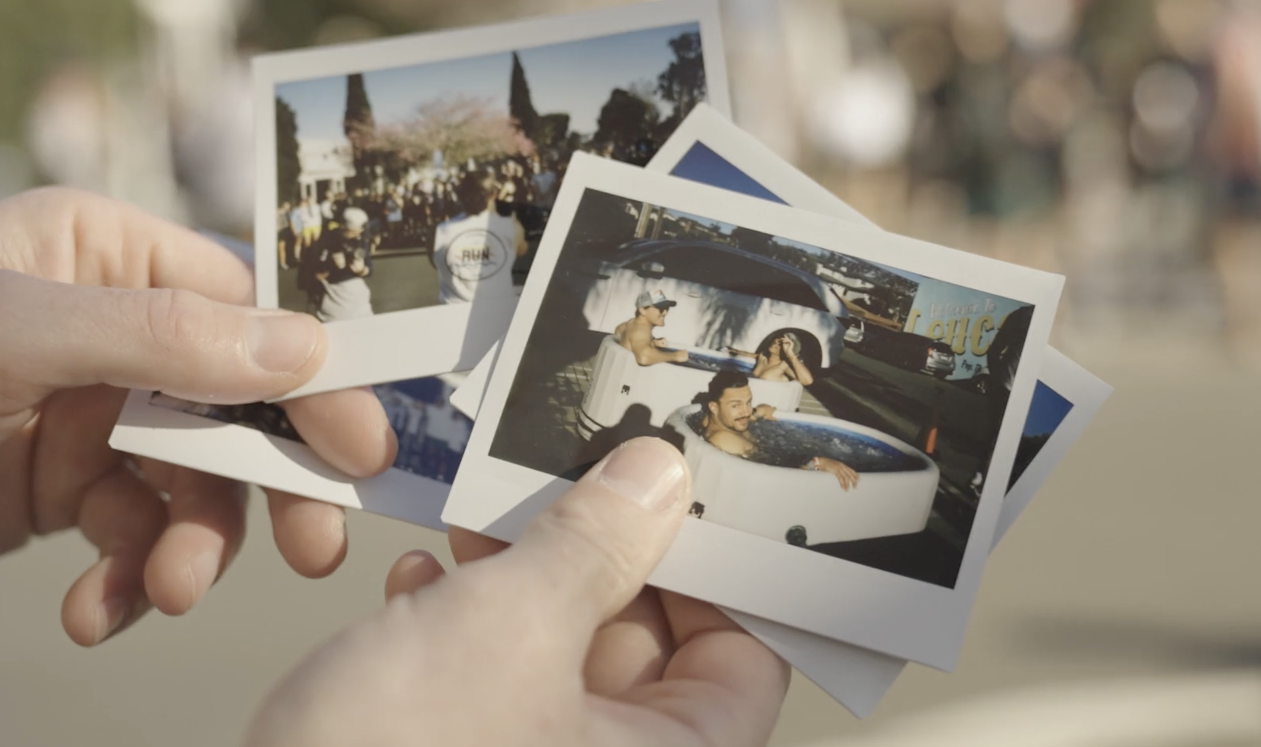 Close-up of a hand holding several printed photographs, one showing a group of people gathered outdoors and another with people in a hot tub, with a white vehicle in the background.