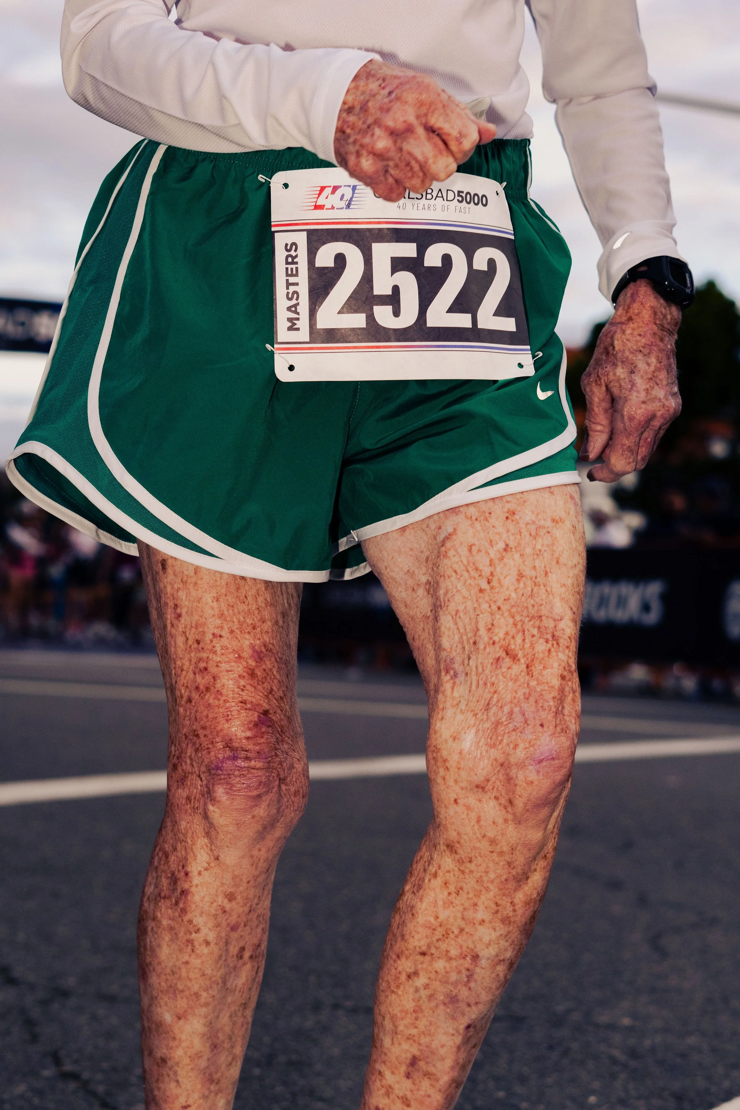 Close-up of a runner's lower body during a race, wearing green shorts and a white long-sleeve shirt, with a race bib numbered 2522 attached to the shorts. The runner's skin appears freckled or sun damaged, and they are wearing a black watch on the le