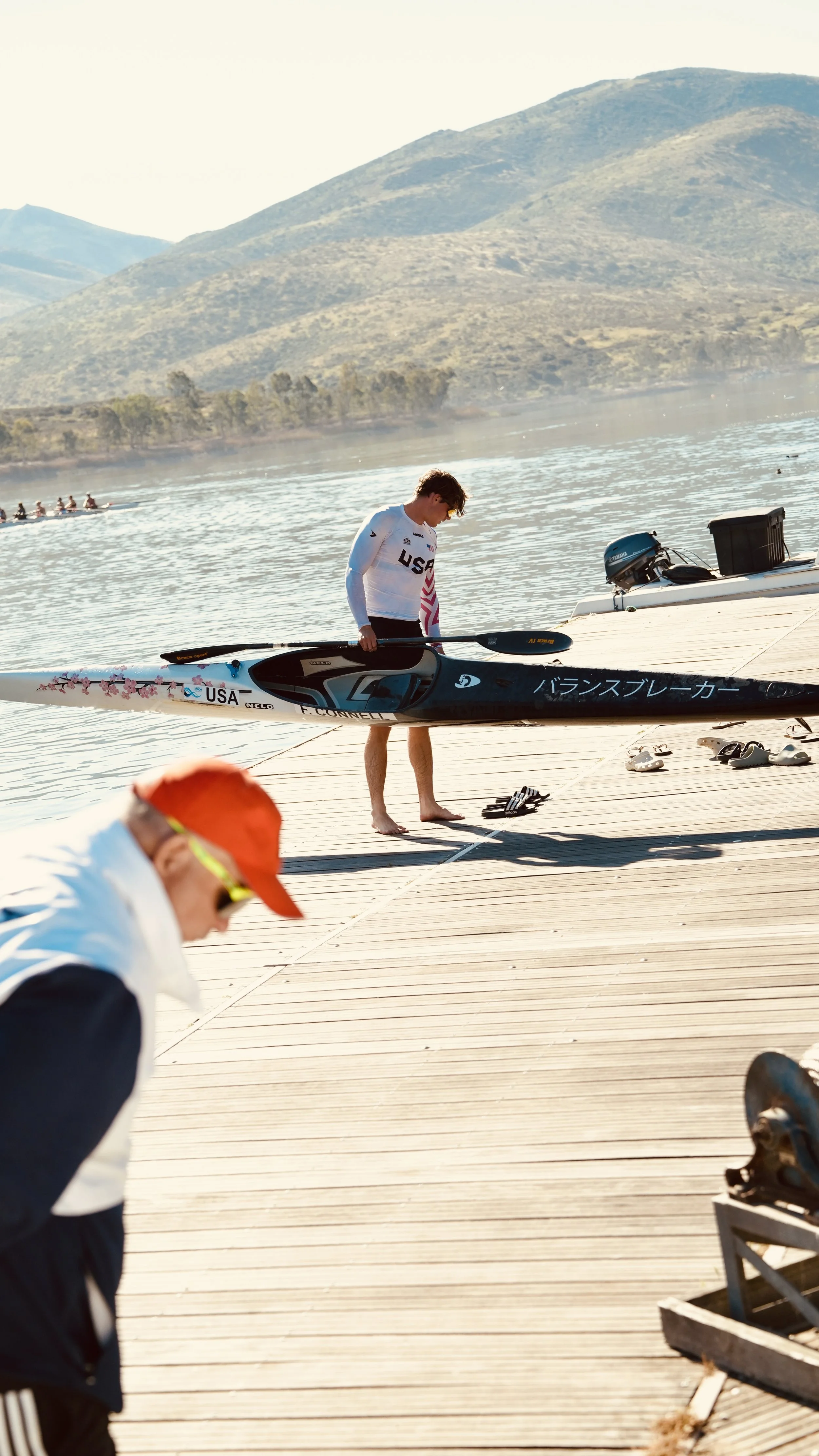 A person in a white USA rowing uniform holding a boat paddle, standing on a wooden dock near a body of water with mountains in the background.