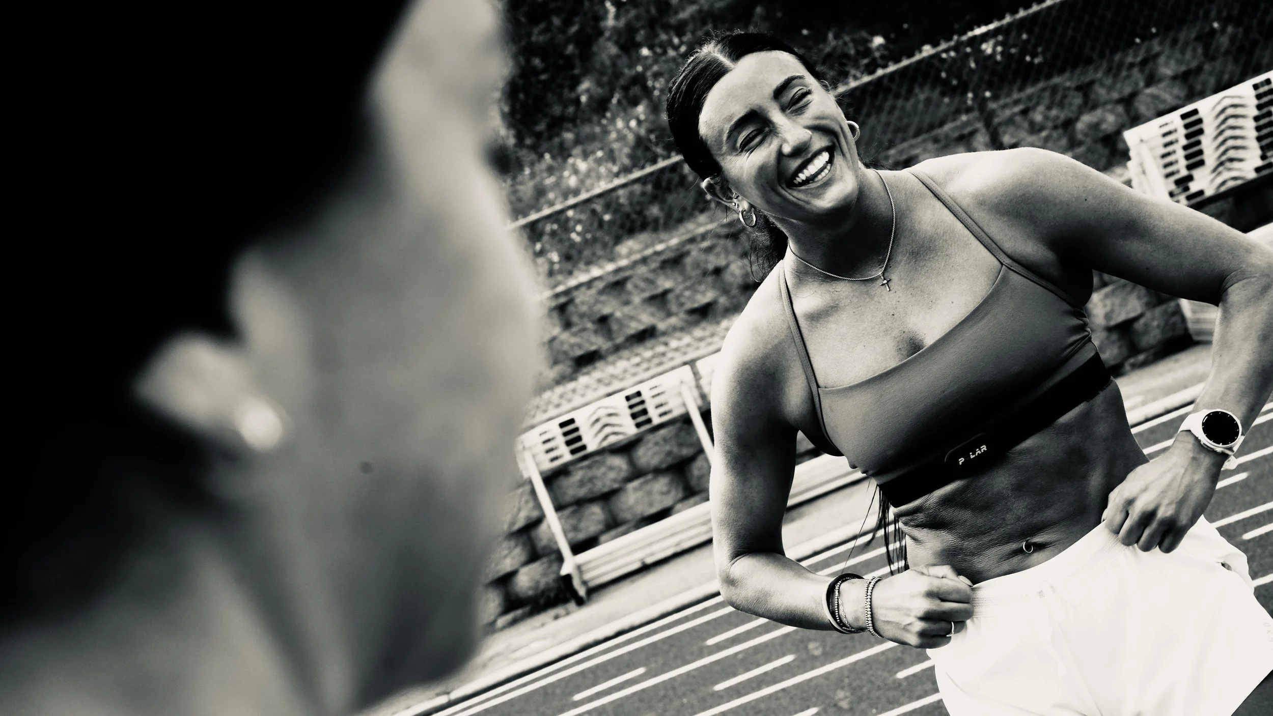 Black and white photo of a smiling woman in athletic clothing, standing outdoors near a bench and a brick wall, likely after a workout or run.