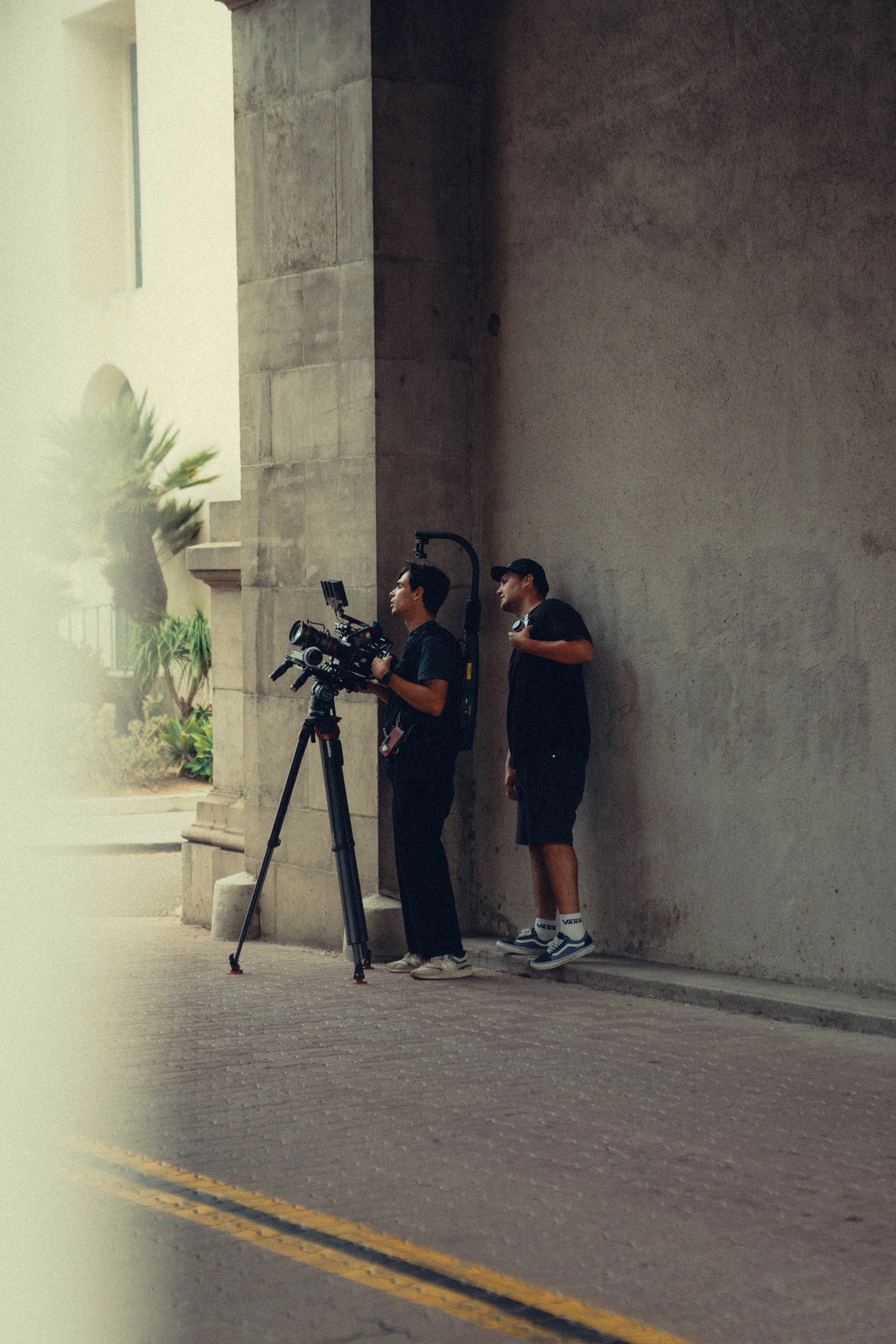 Two young men, one operating a professional camera on a tripod and both standing against a large stone wall, appear to be filming or photographing. The scene is outdoors with some greenery and a building in the background, possibly on a city street.