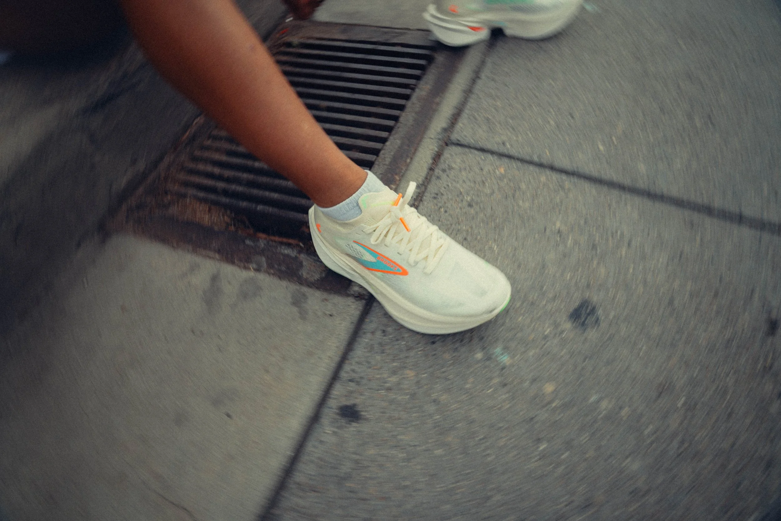 Close-up of a person's foot wearing a white sneaker with orange and blue accents, on a concrete sidewalk near a metal street drain grate. The person is wearing light-colored socks and a short-sleeved shirt.