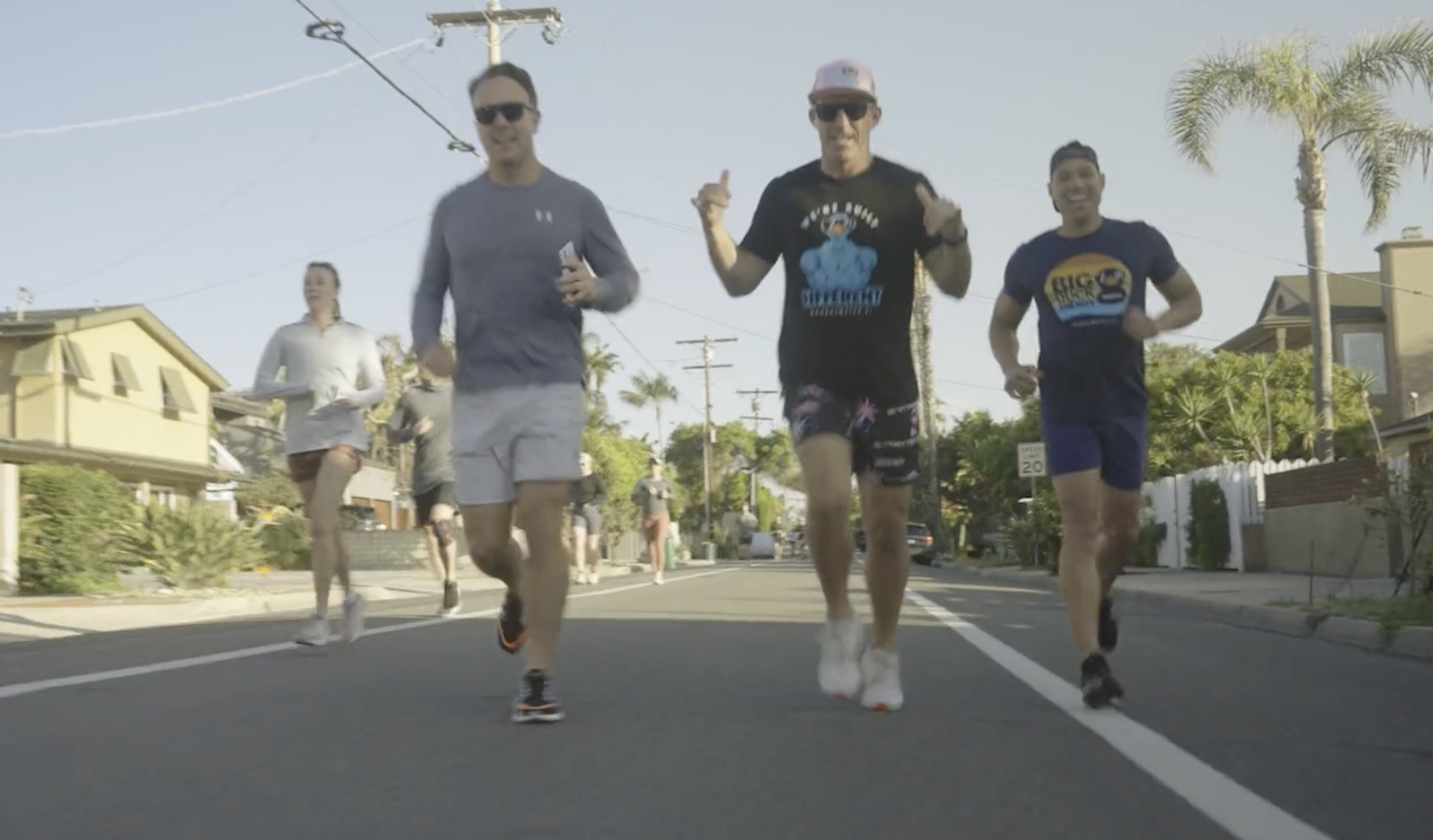 Group of five runners jogging on a residential street during daytime, wearing casual athletic clothing, with palm trees and houses in the background.