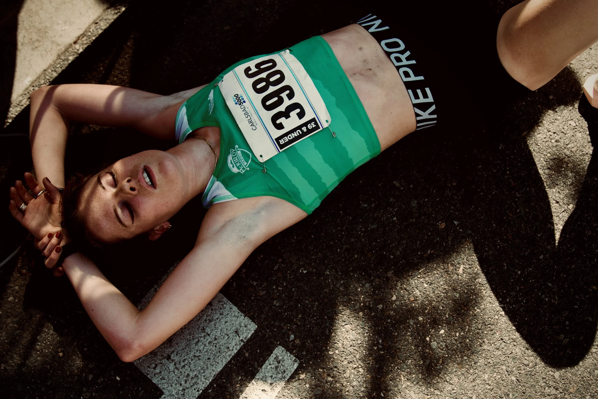 A woman lying on the ground on a running track, wearing a green sports outfit with a bib number 3986. She appears to be tired or resting after a race.