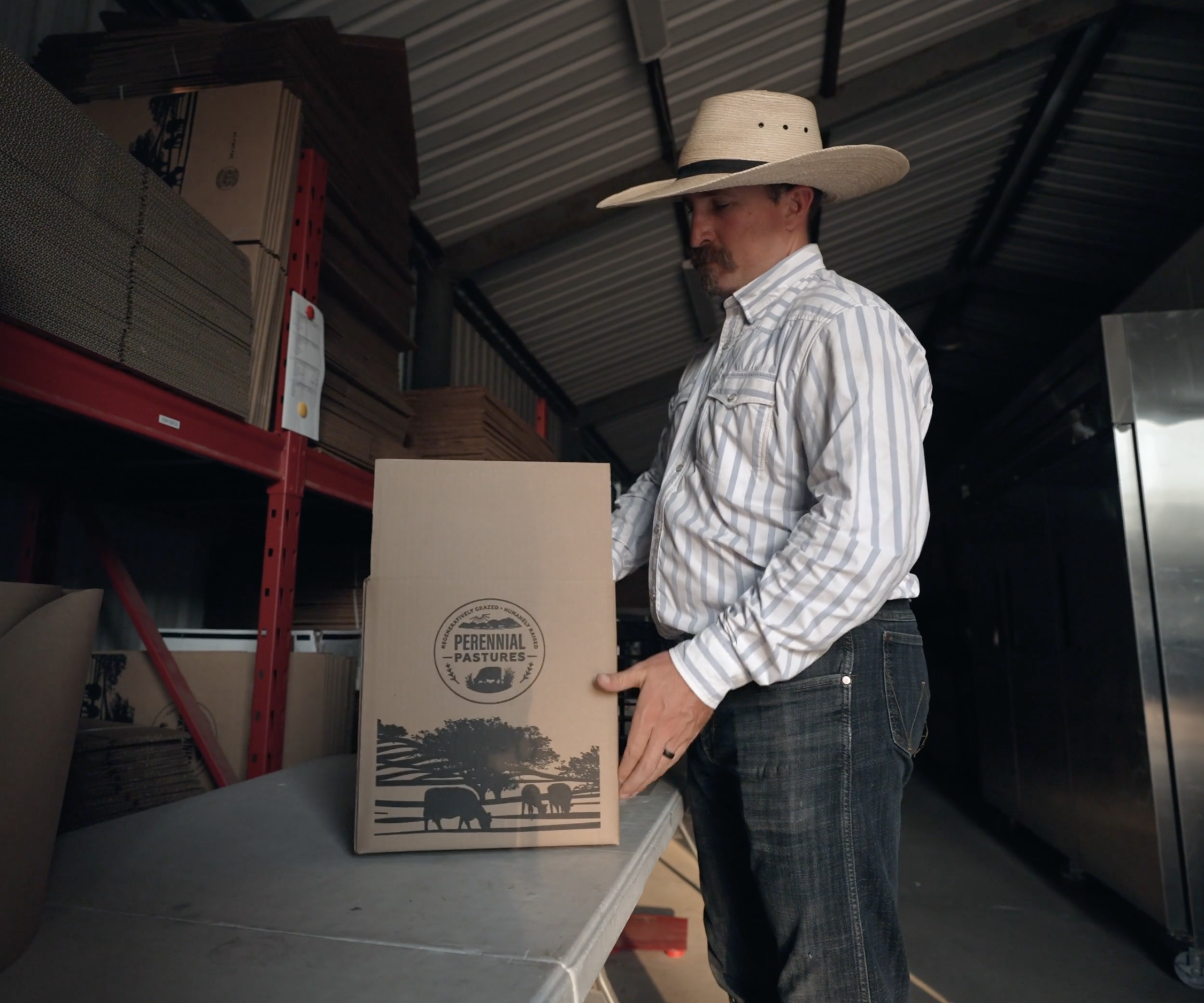 A man with a beard wearing a wide-brimmed straw hat and a striped shirt shops for boxes on metal shelves in a warehouse, holding a cardboard box labeled "Perennial Pastures."