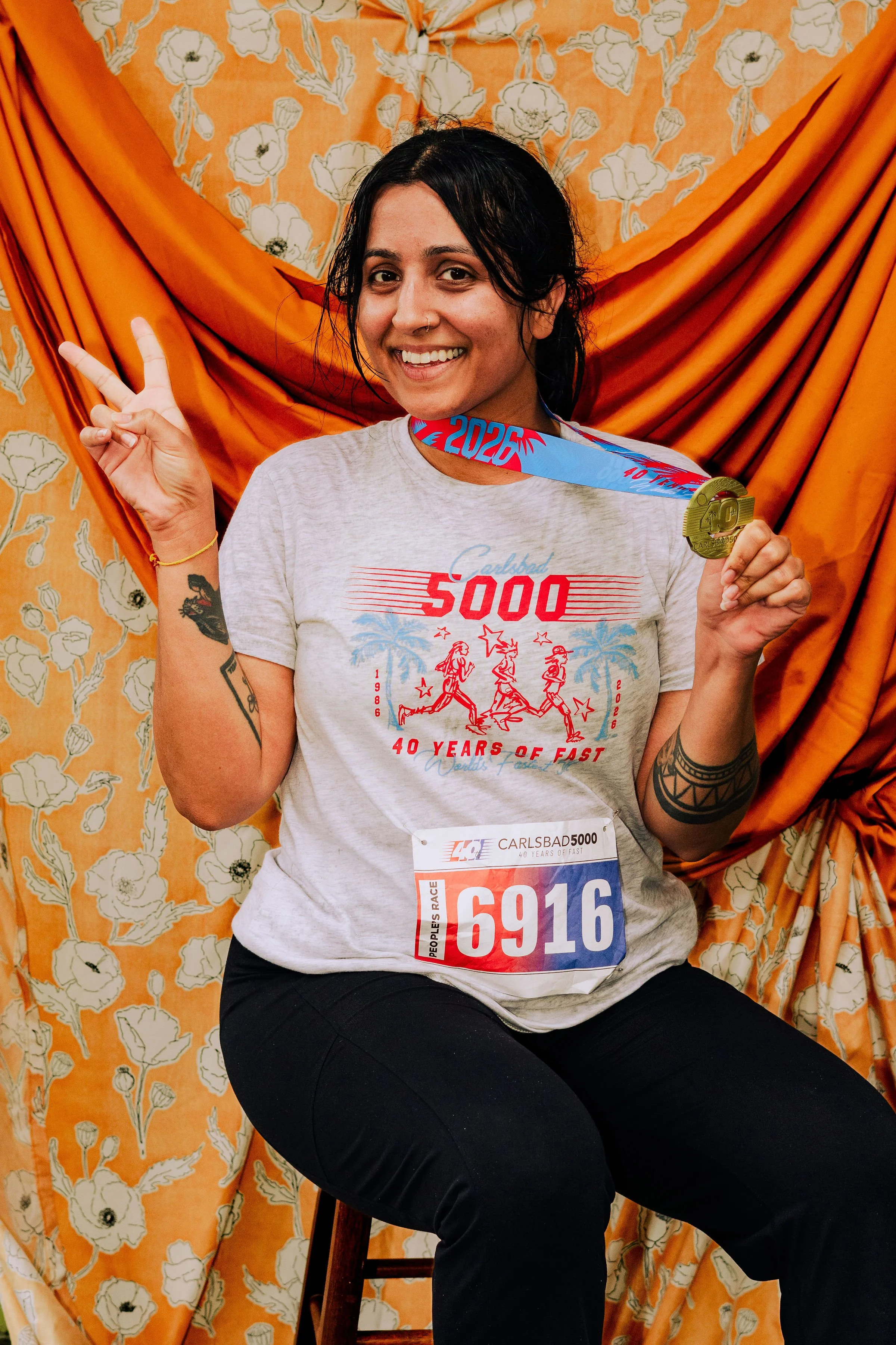 A smiling woman with black hair and tattoos, wearing a t-shirt and a race bib, seated on a wooden chair in front of a floral orange and cream curtain backdrop. She is holding a medal and making a peace sign with her right hand.