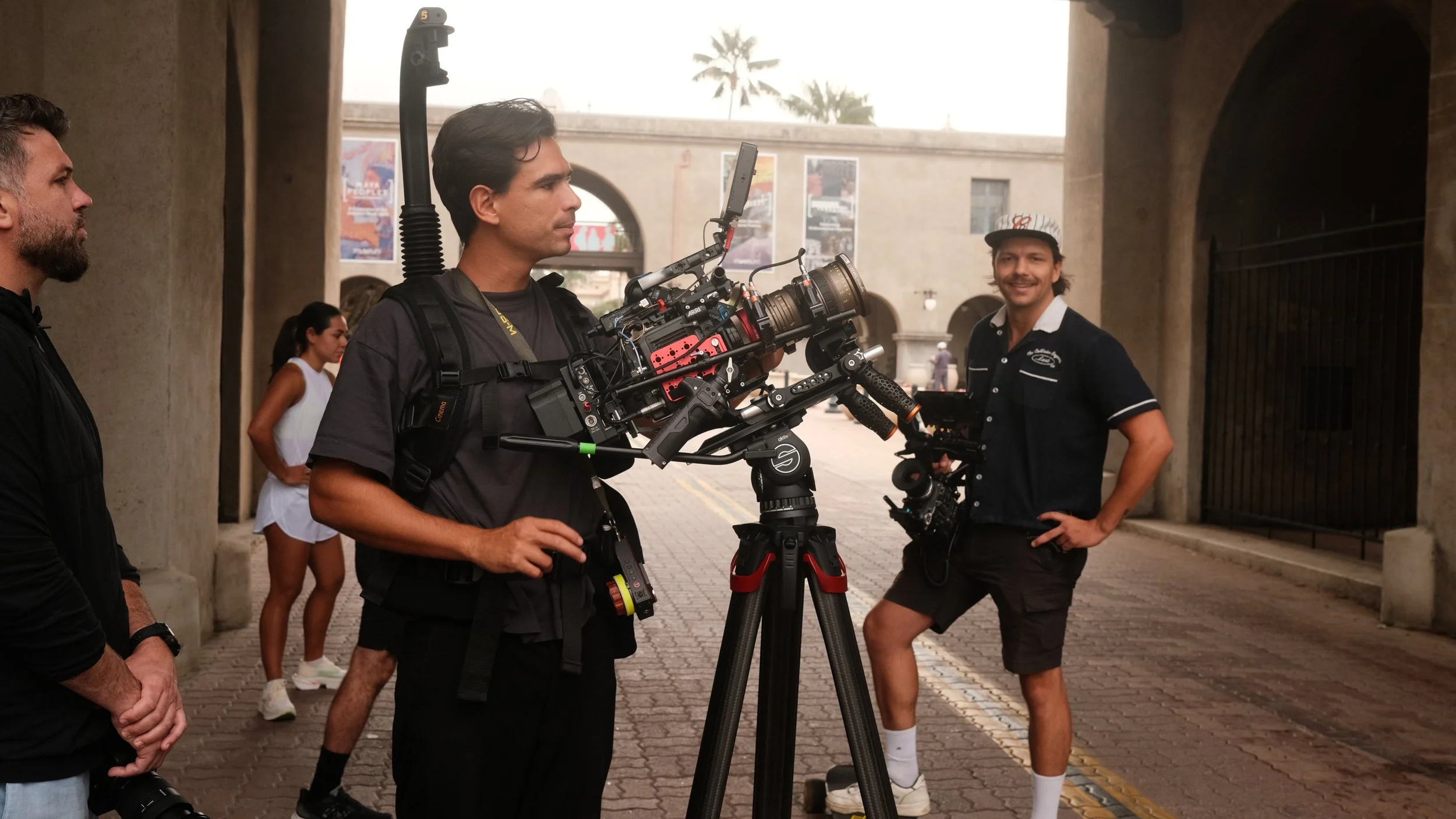 Film crew setting up camera equipment outdoors with a man smiling in the background.