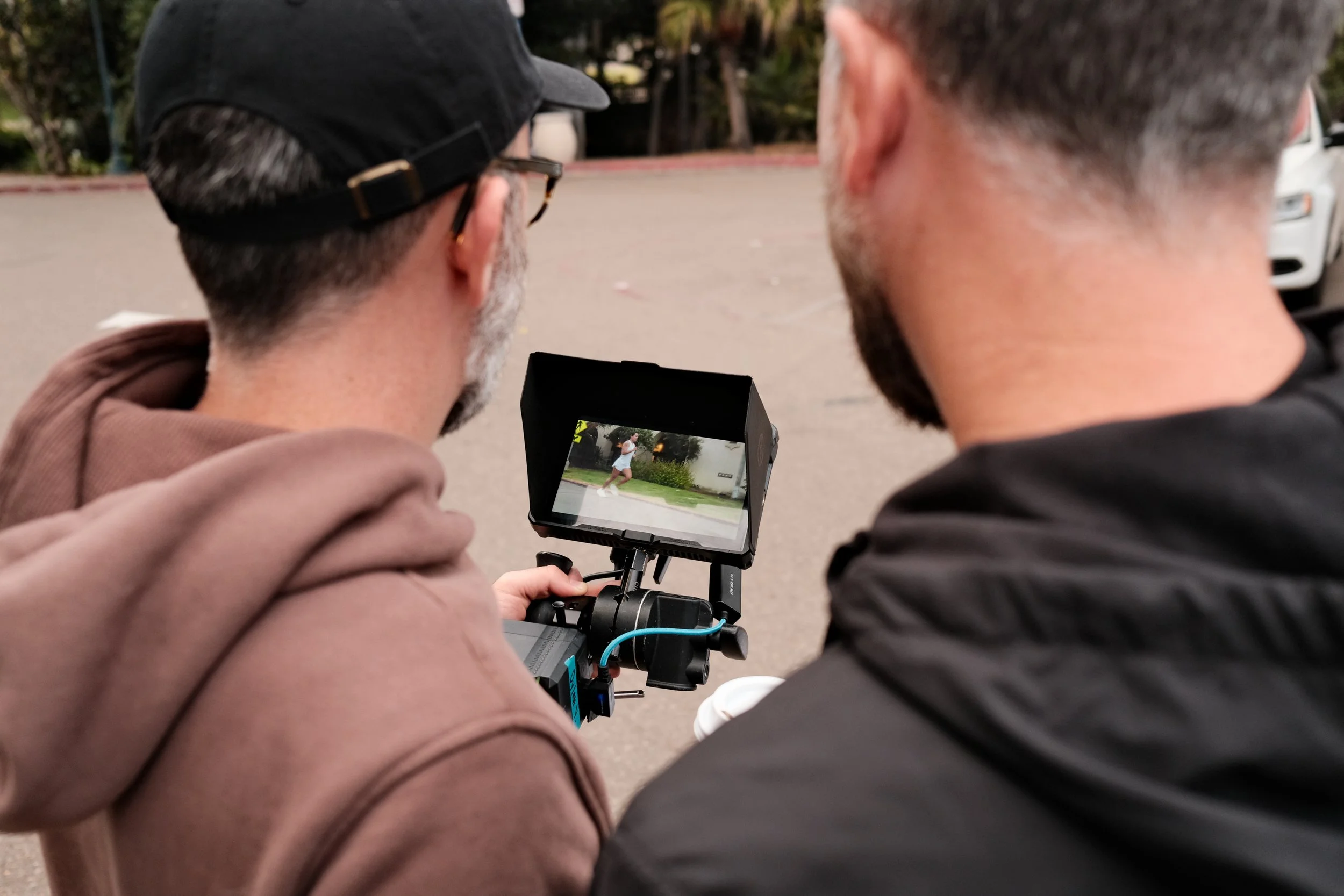 Two men stand close together outdoors, one man is wearing a baseball cap and glasses, and the other has short hair and a beard. They are looking at a camera or monitor, which displays a picture of a person running.