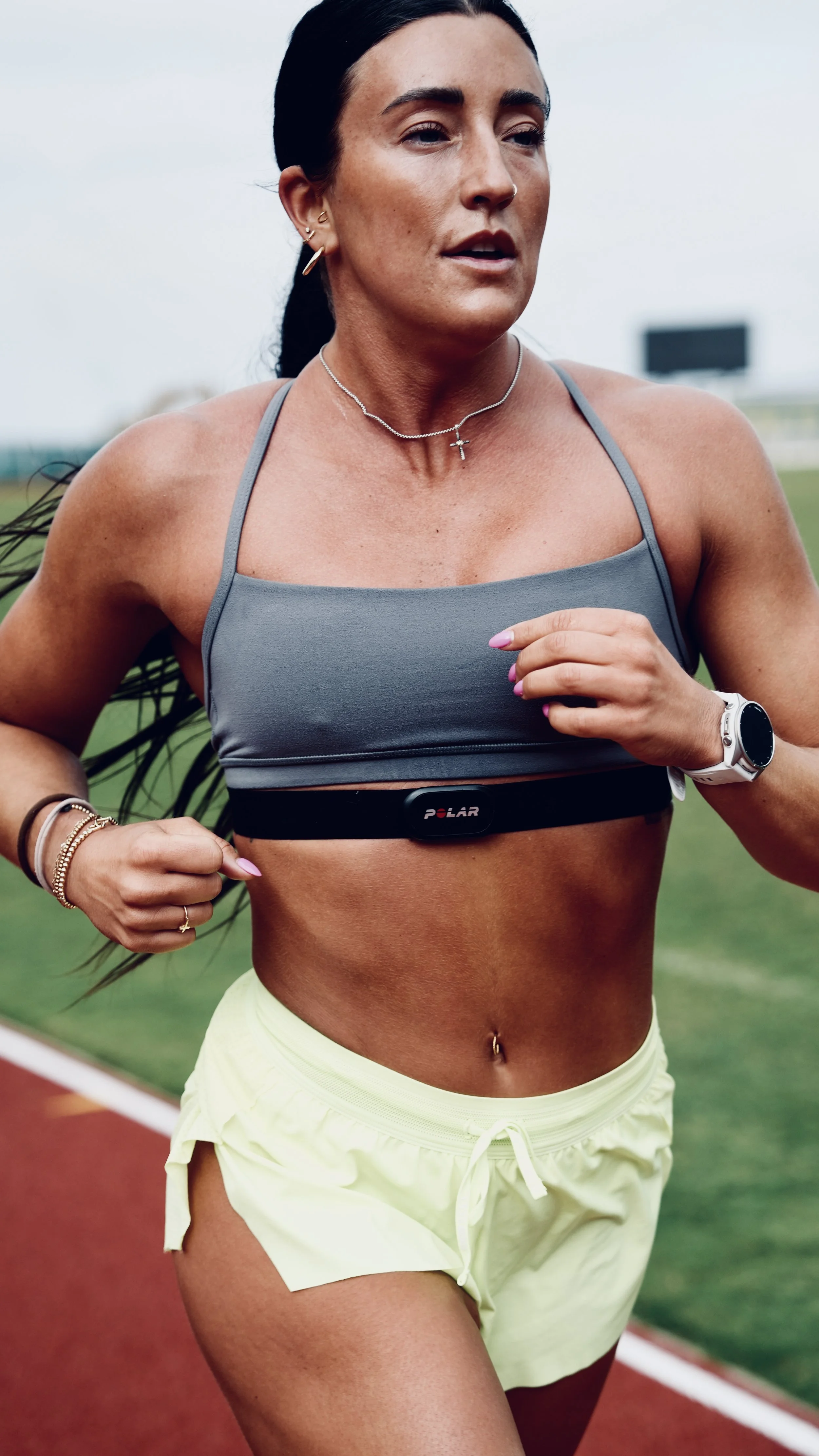 A woman running on a track wearing a gray sports bra and yellow shorts, with a heart rate monitor around her chest, a smartwatch on her left wrist, and jewelry including necklaces and earrings.