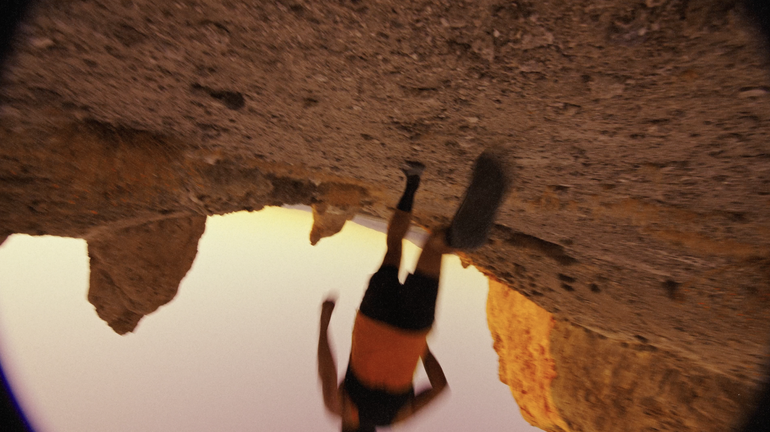 Person rock climbing on a rugged brown rock face during sunset with orange sky.