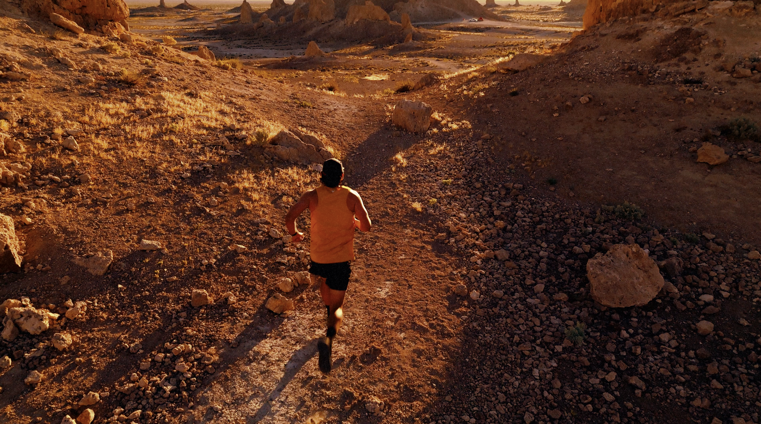 A person running on a rocky trail through a desert landscape at sunset with rocky formations in the background.