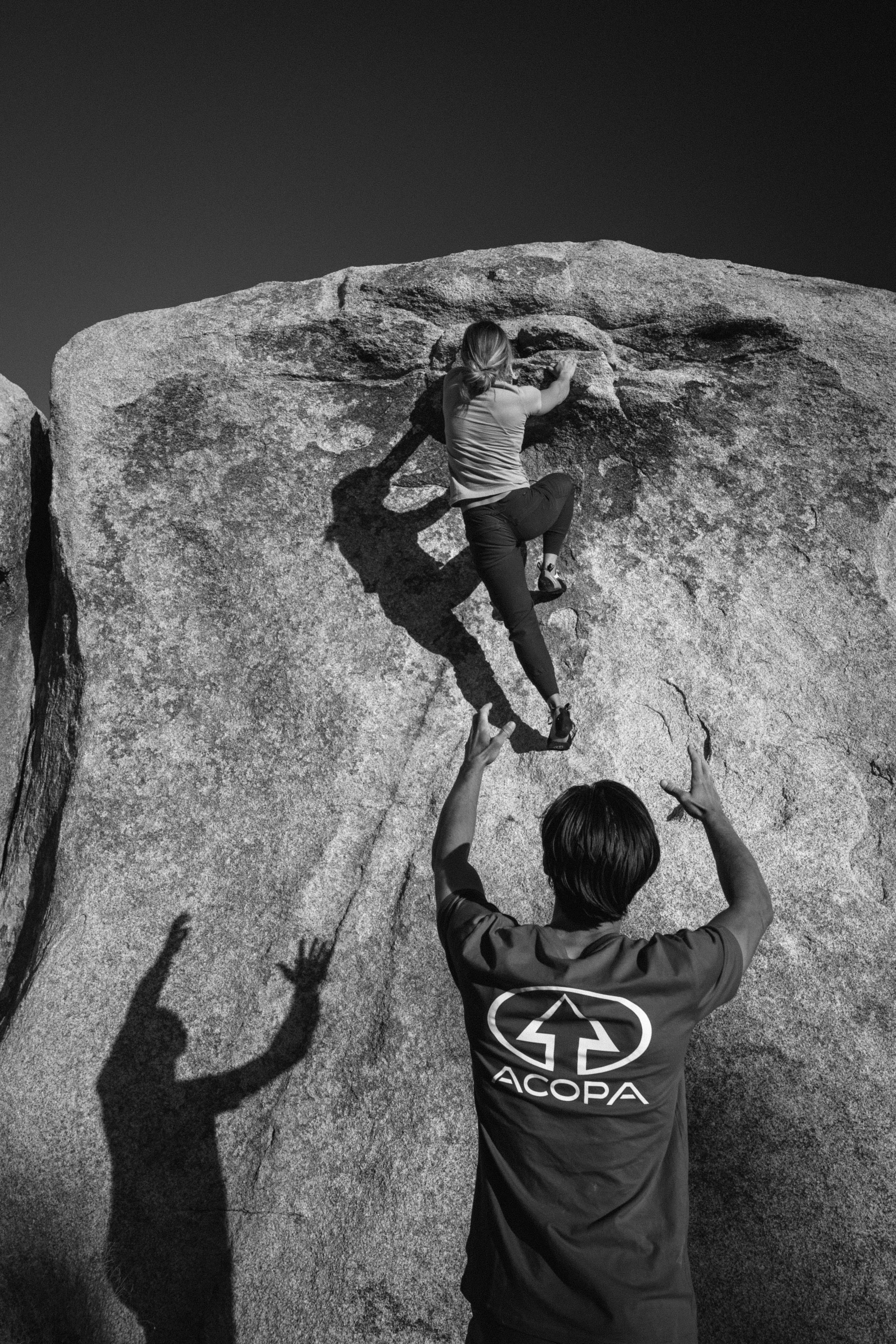 A woman rock climbing on a large granite boulder outdoors, with two people below spotting her.