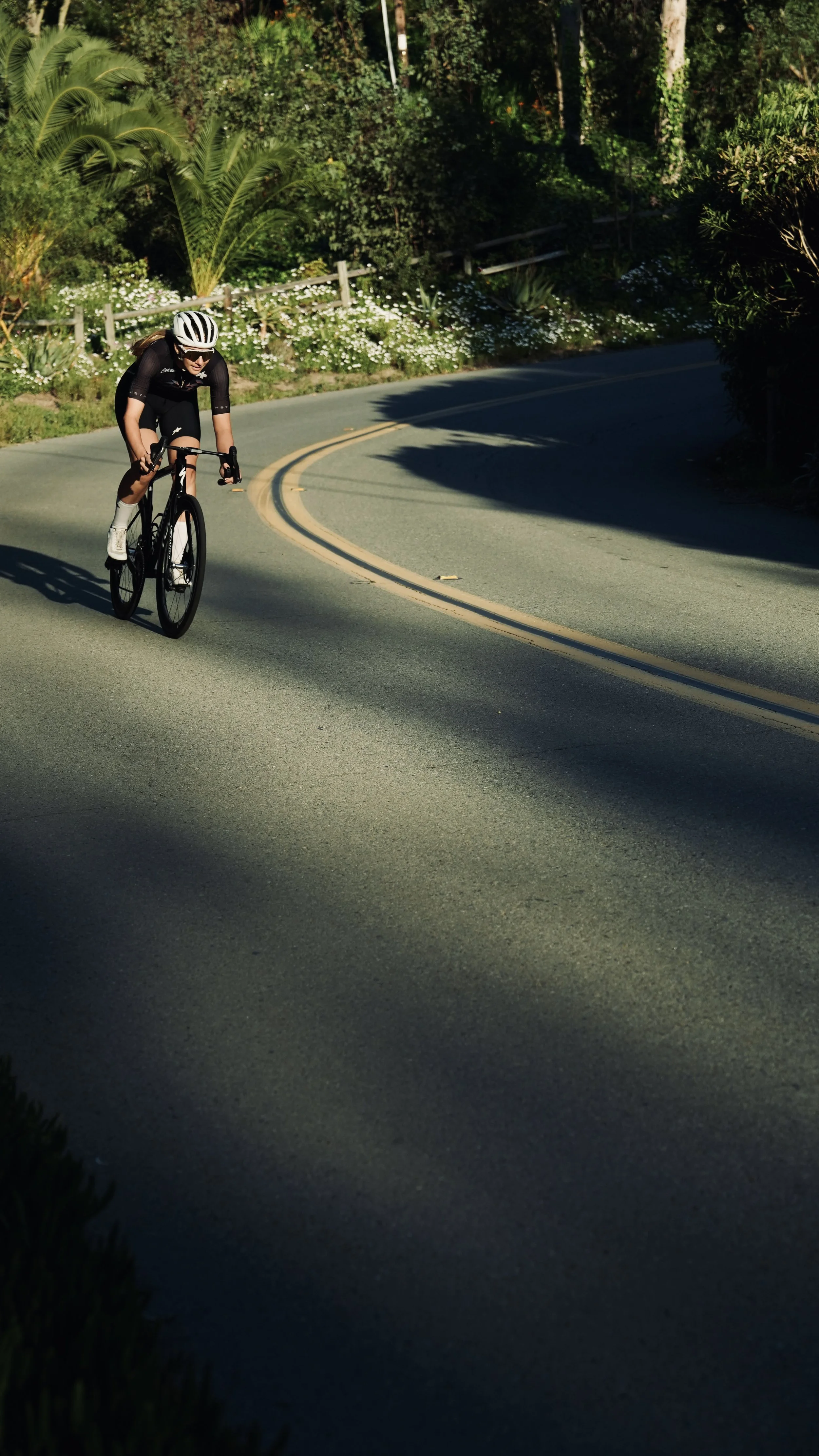A woman in black cycling gear and a white helmet riding a bicycle along a winding road surrounded by lush green trees and bushes.
