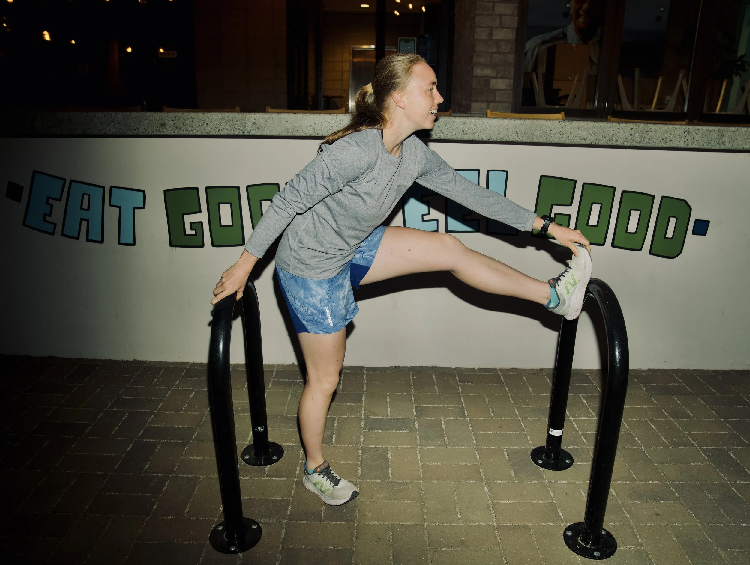 Young woman stretching outside at night, on a stretch machine, wearing a gray long-sleeve shirt, blue shorts, and athletic shoes, with a smile.