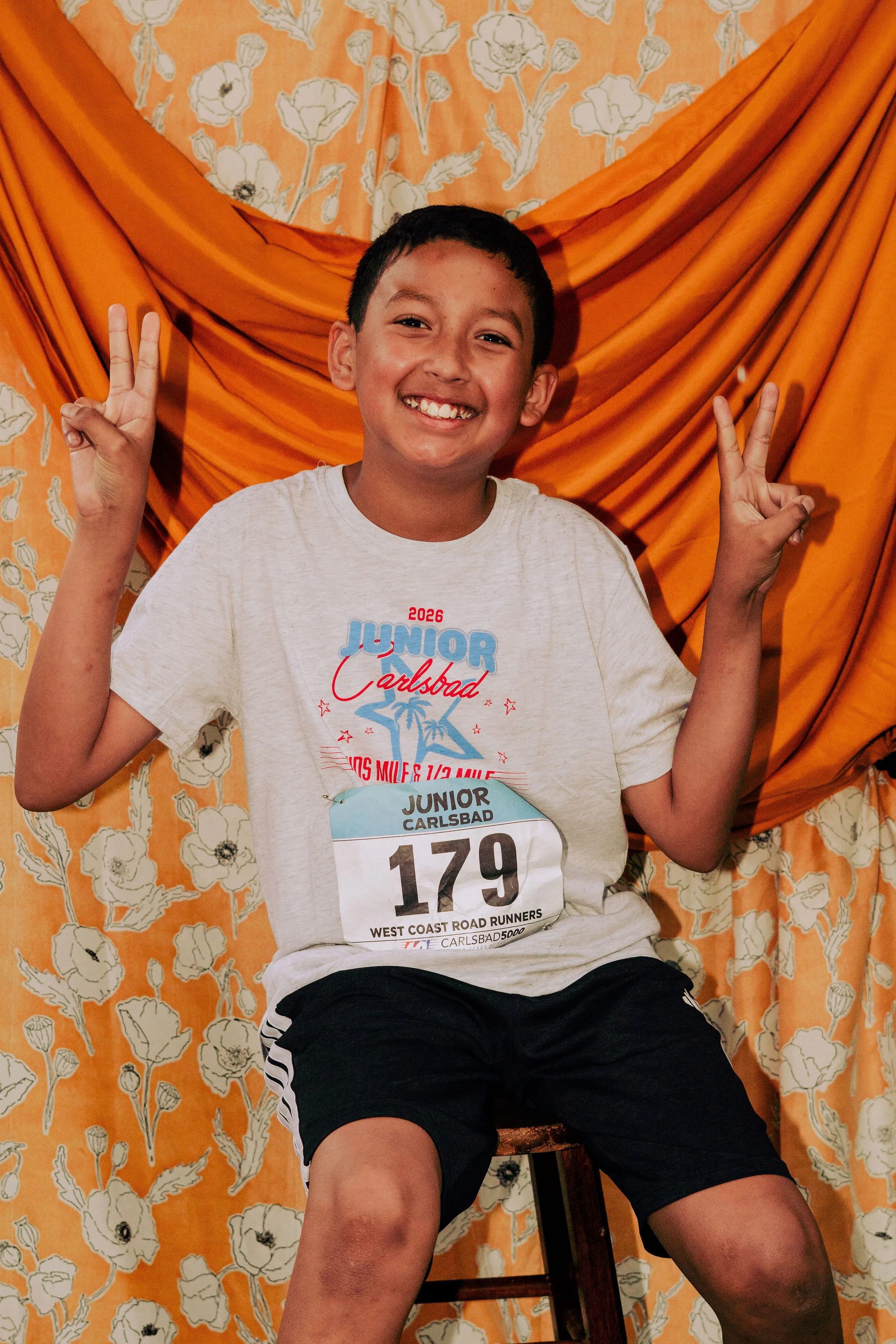 A smiling young boy making peace signs with both hands, sitting on a stool in front of an orange floral curtain, wearing a race bib for a running event.