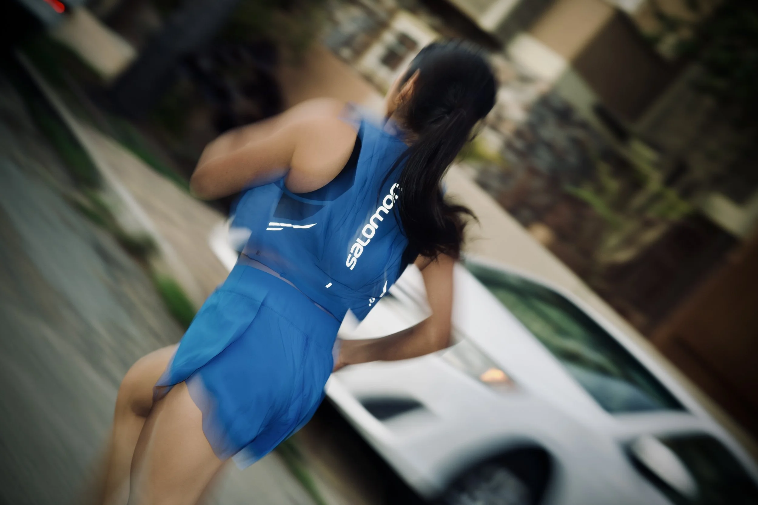 A female athlete from behind, wearing a blue sports uniform with 'Salomon' written on it, with long black hair tied back, bending forward with her hands on her knees, on an indoor sports court.