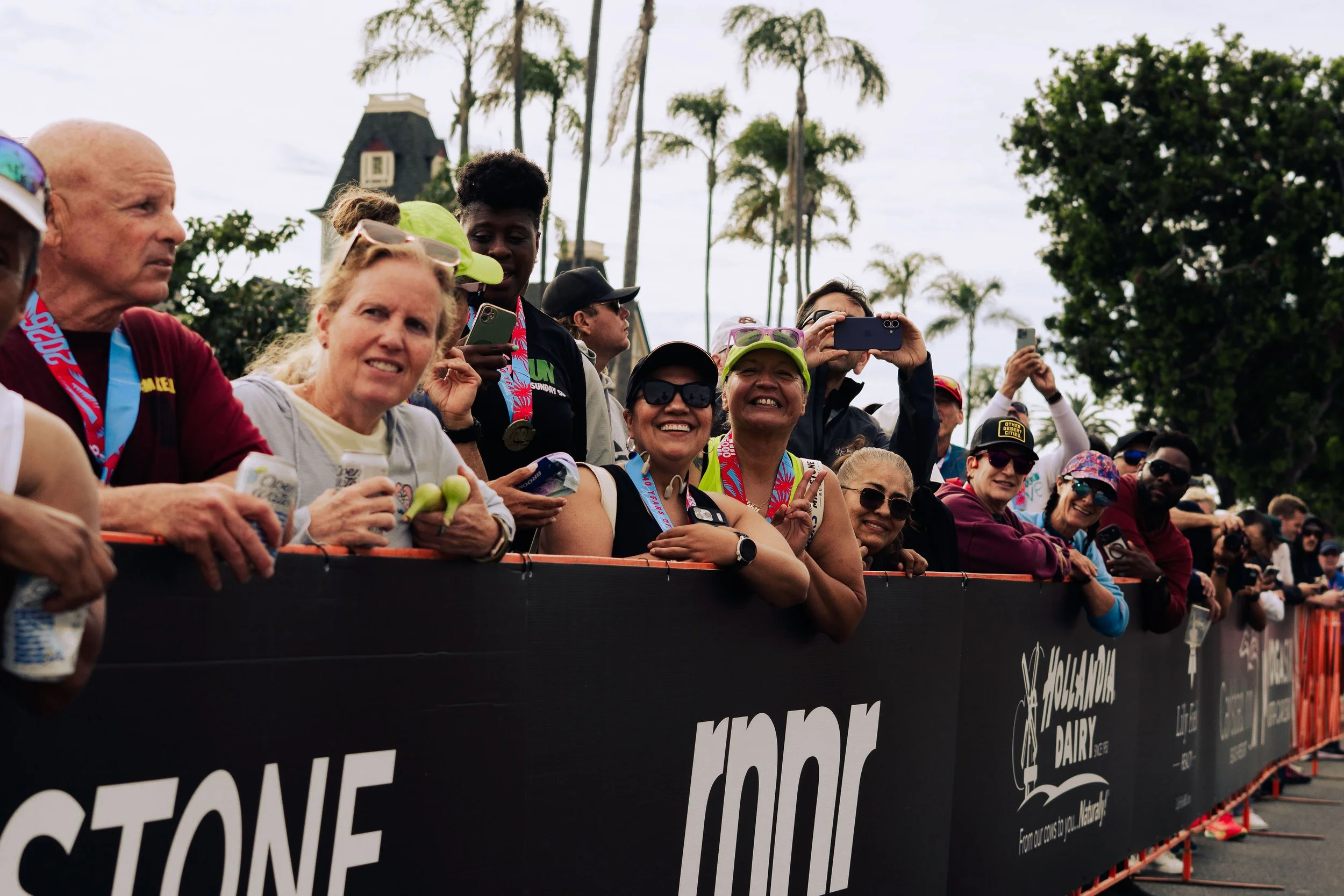 Group of people standing behind a barricade, smiling and taking photos at an outdoor event under cloudy skies, with trees in the background.