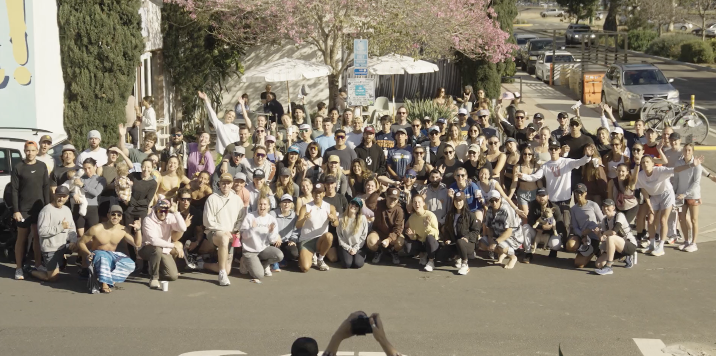 A large group of people gathered outdoors in a parking lot on a sunny day, some waving or making hand gestures, with trees, parked cars, and umbrellas in the background.