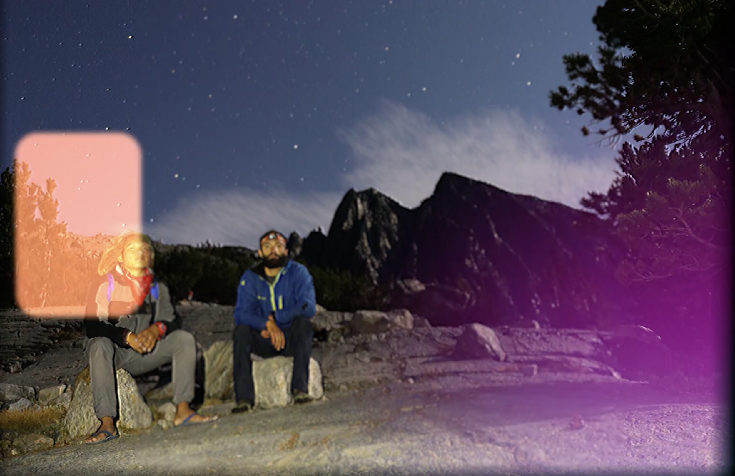Two hikers sitting on rocks against a starry night sky in a mountain area, with trees and mountain peaks visible.