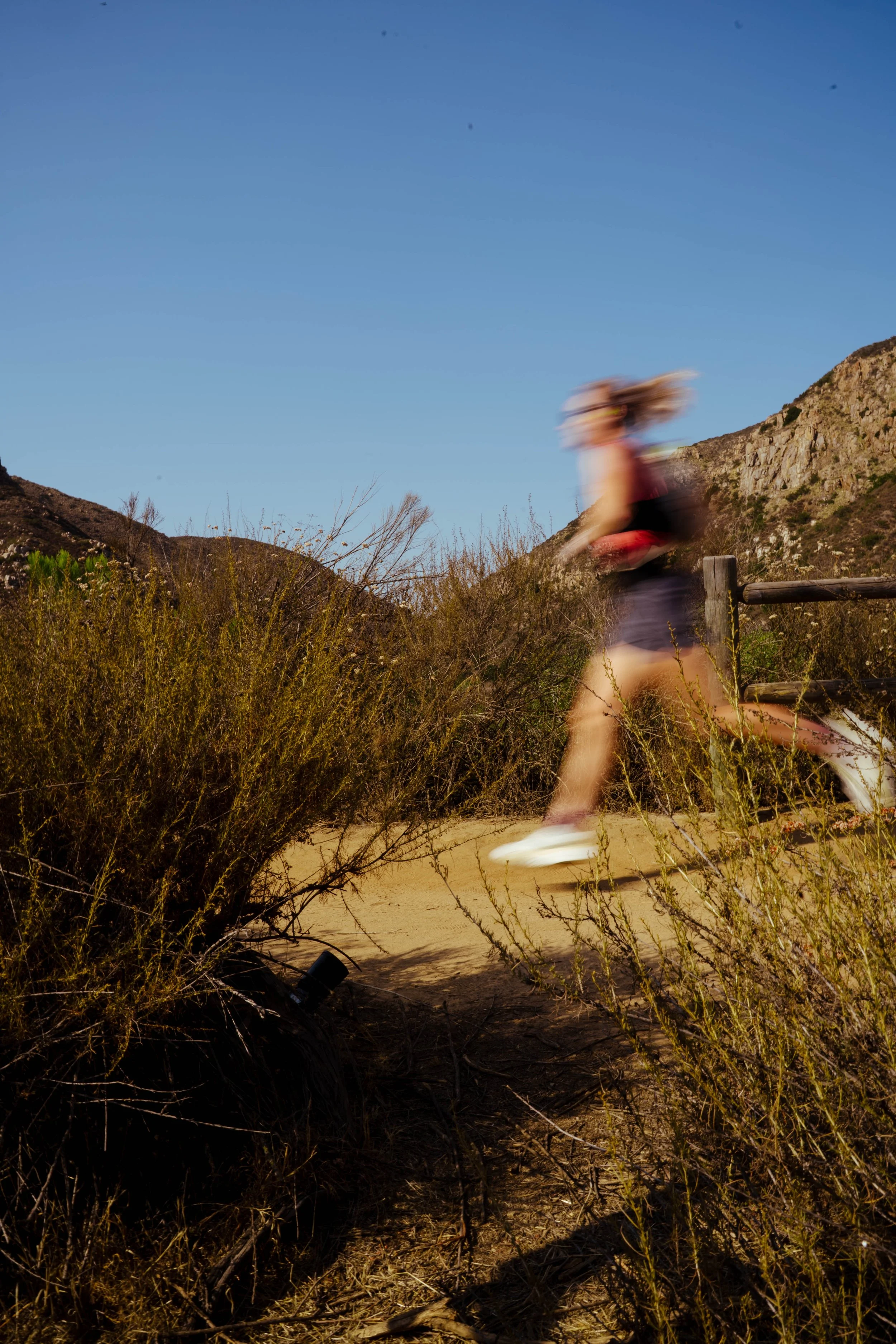A woman running outdoors on a trail with hills and bushes, blurred due to motion, in a sunny, natural environment.