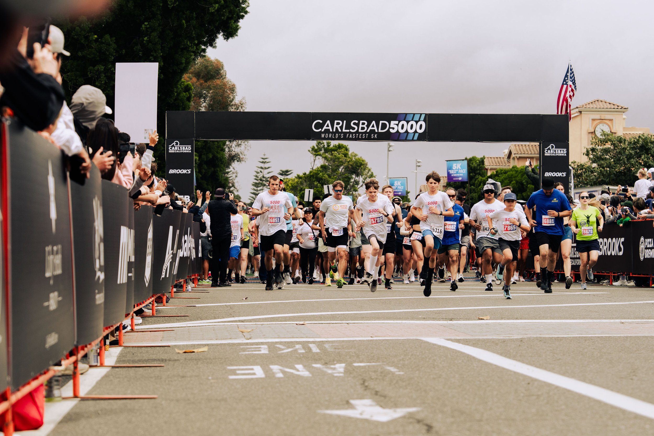 Runners competing in a marathon race at the starting line with a black finish line banner that reads 'Carlsbad 5000 World's Fastest 5K' and spectators cheering on the sides.