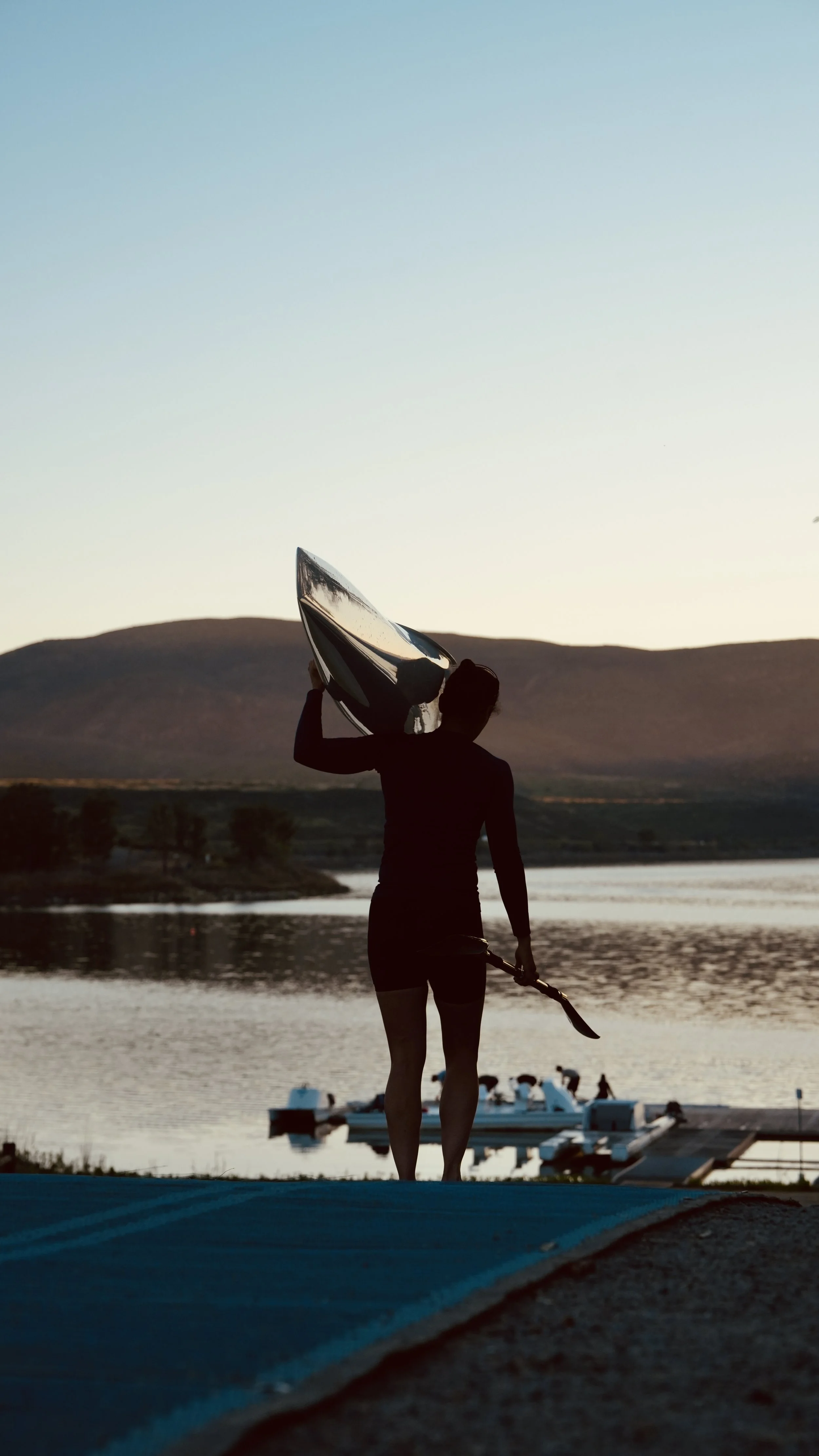 Silhouette of a woman holding a paddle and a kayak paddle, standing near a lake during sunset, with mountains in the background and a boat docked nearby.