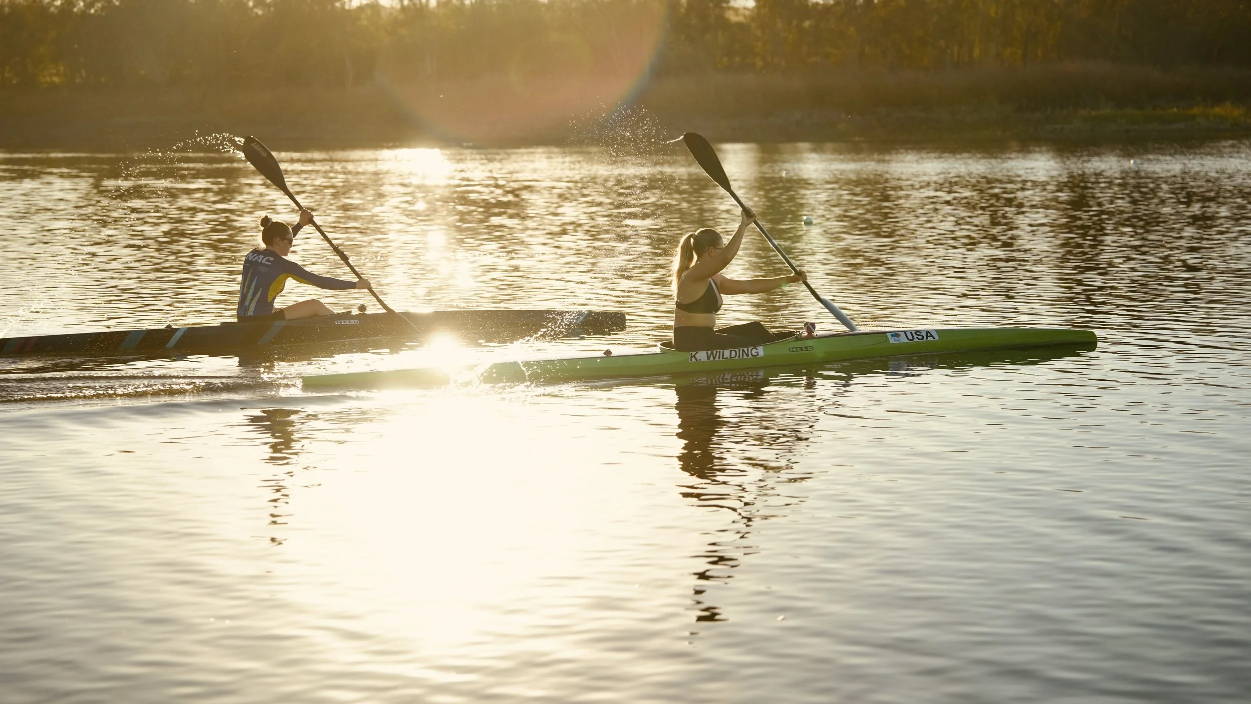 Two women paddleboarding on a calm body of water at sunset, with the sun casting a warm glow and reflections on the water surface.