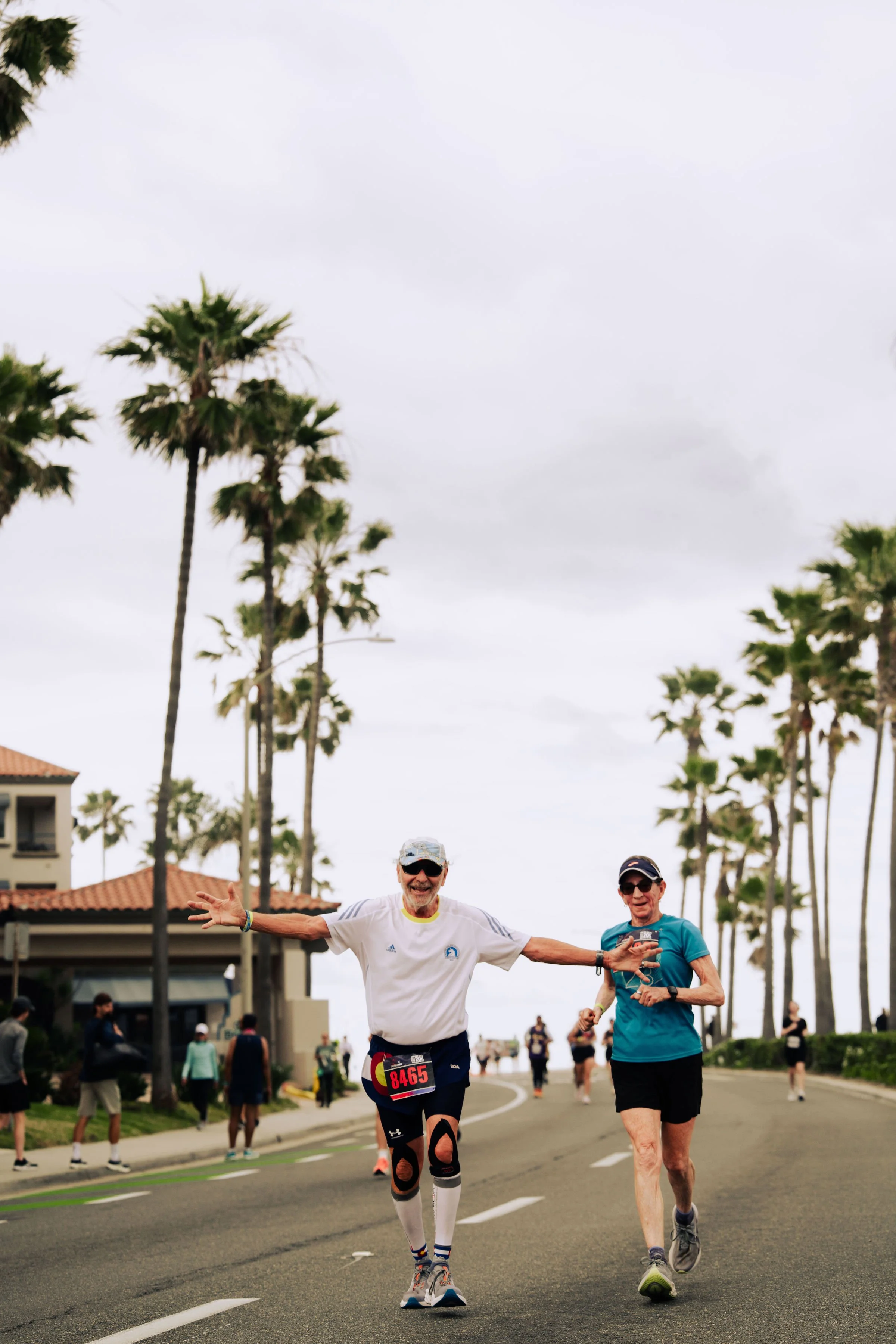 Two elderly runners celebrating during a race on a street lined with palm trees, with participants and spectators in the background.