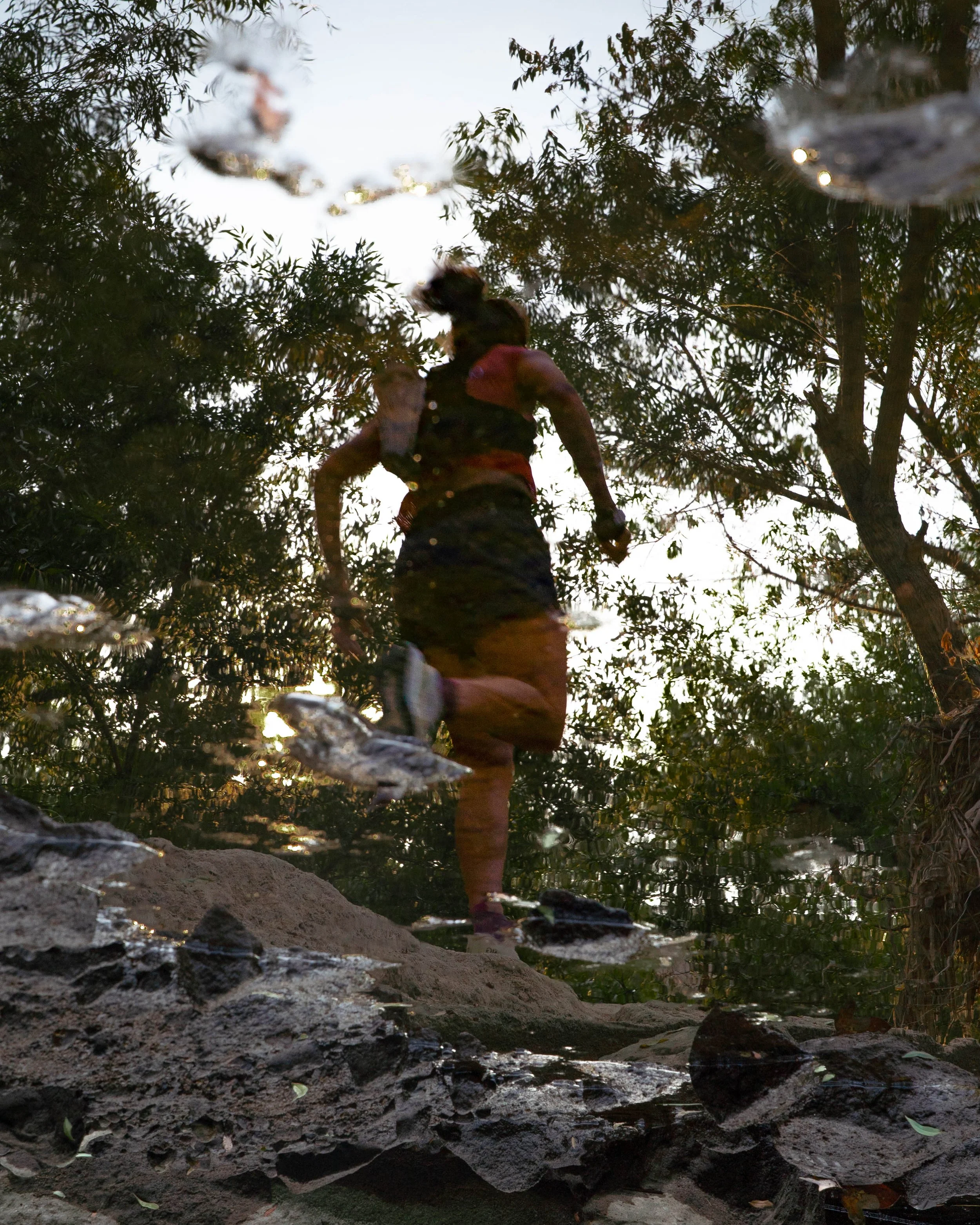 A person running through a forested trail with trees on both sides, seen from a low angle near the ground, with water and rocks in the foreground, and daytime sunlight filtering through the branches.