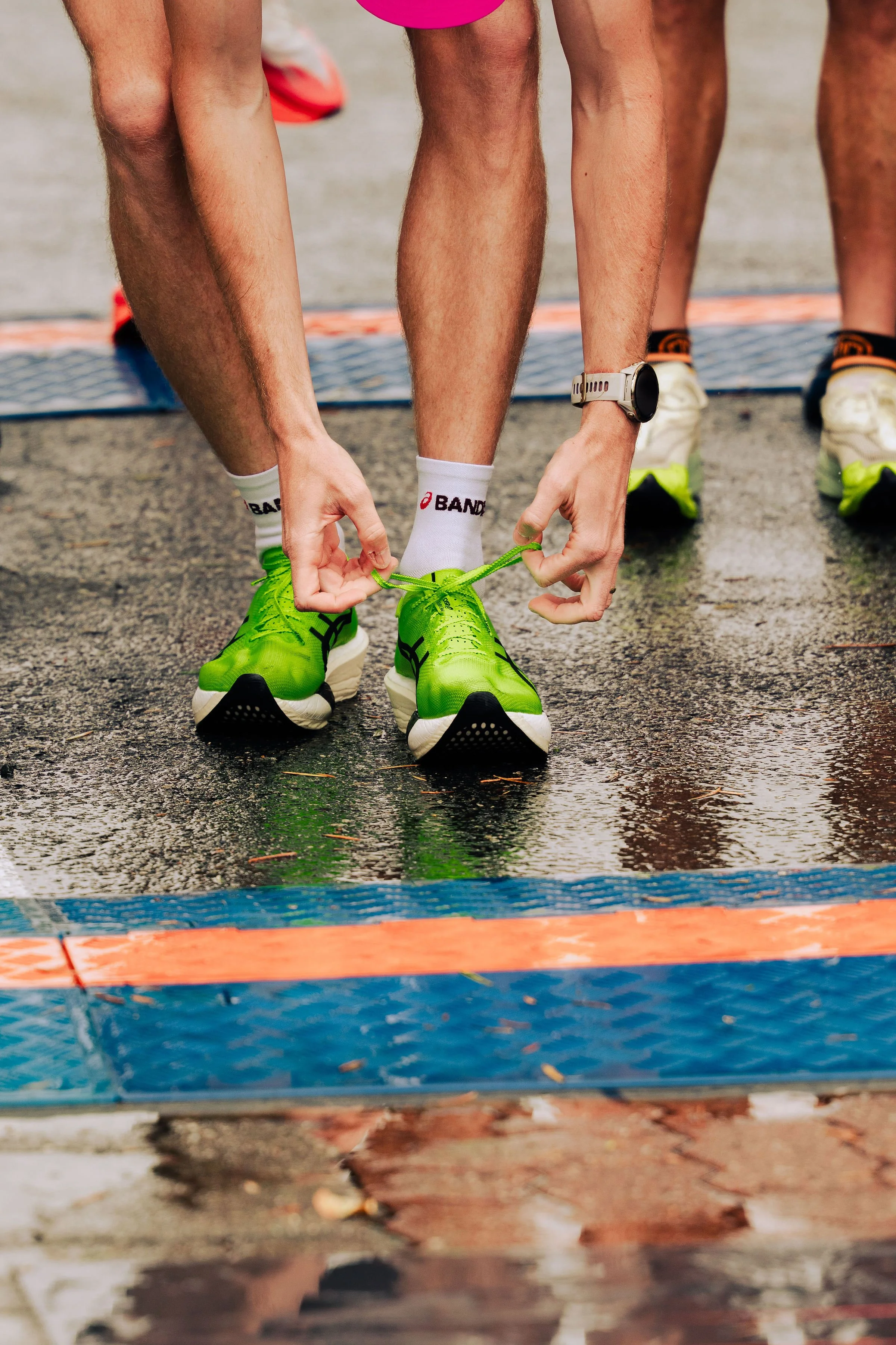 Close-up of a runner tying shoelaces on bright green athletic shoes at a race starting line, with other runners' legs visible in the background.
