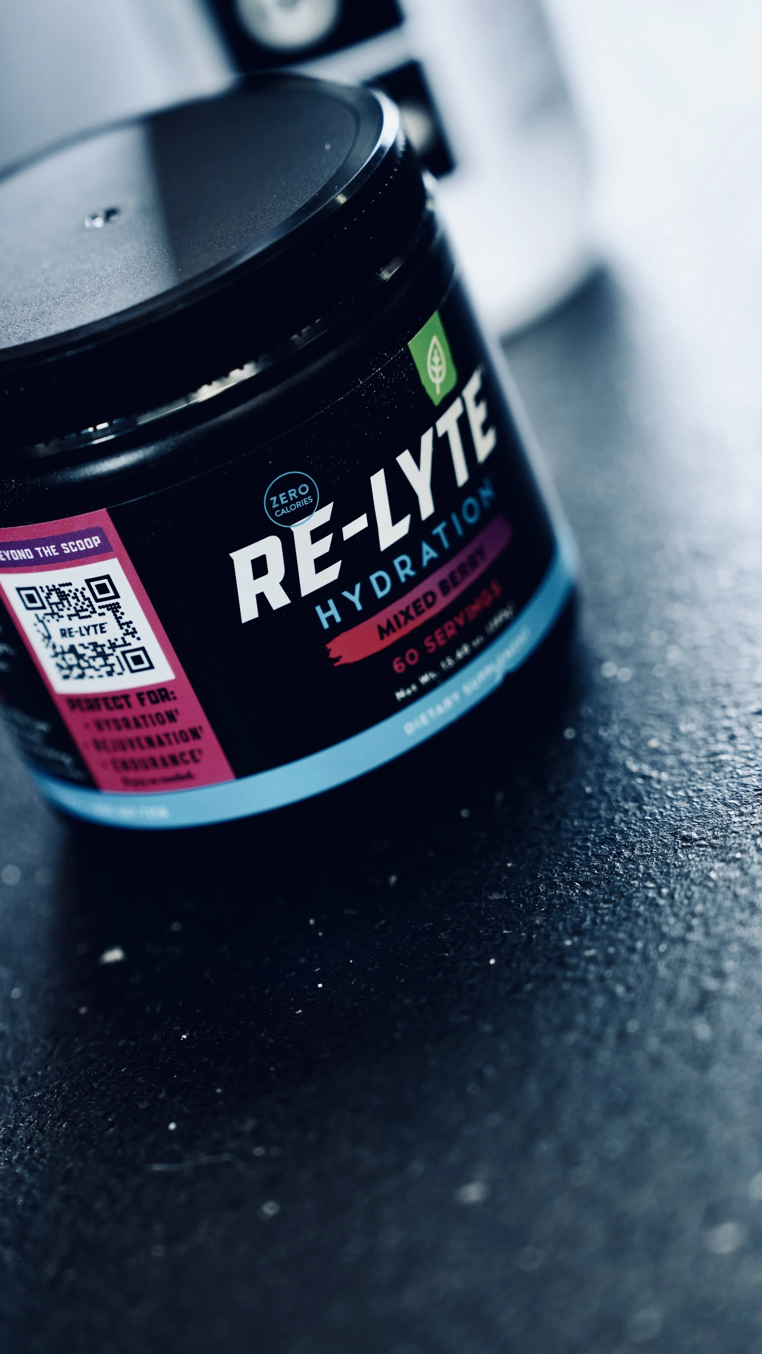 Close-up of a black container of Re-LYTE hydration powder with a green leaf logo, pink and blue accents, placed on a dark surface.