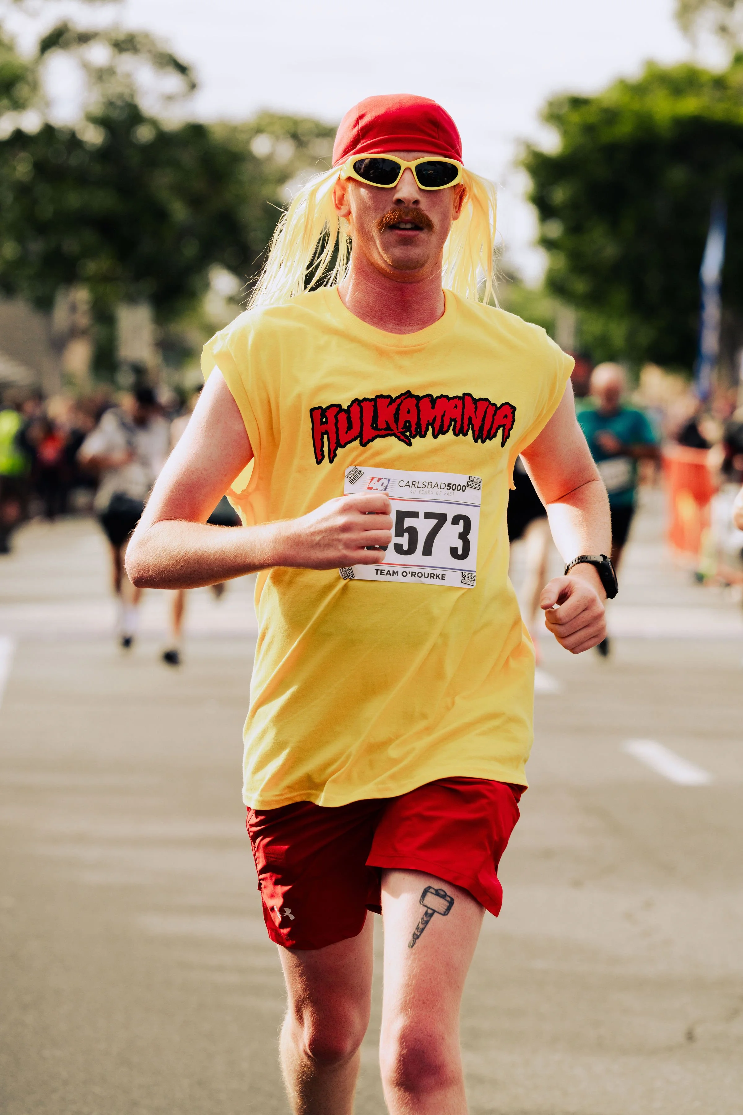 A man running in a race, wearing a yellow Hugo Kwamya shirt, red shorts, a red cap, yellow sunglasses, and a red wristwatch, with a tattoo of a hammer on his left thigh, in an outdoor race on a sunny day with spectators in the background.