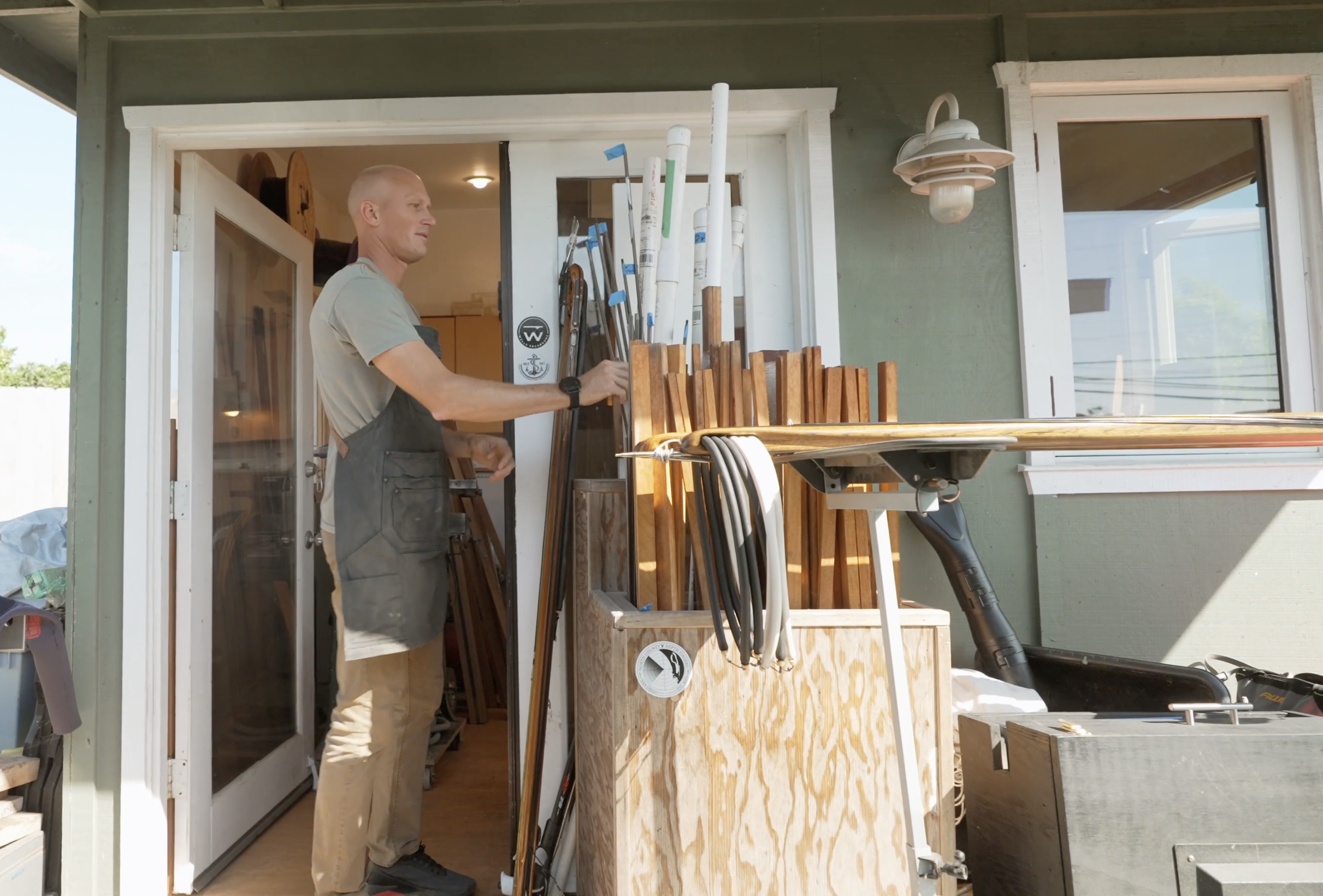A man wearing a gray t-shirt, khaki pants, and a dark apron is working outside a house, organizing tools on a wooden workbench under a covered porch. The house has green siding, white trim, and a large window. The scene is bright and sunny.