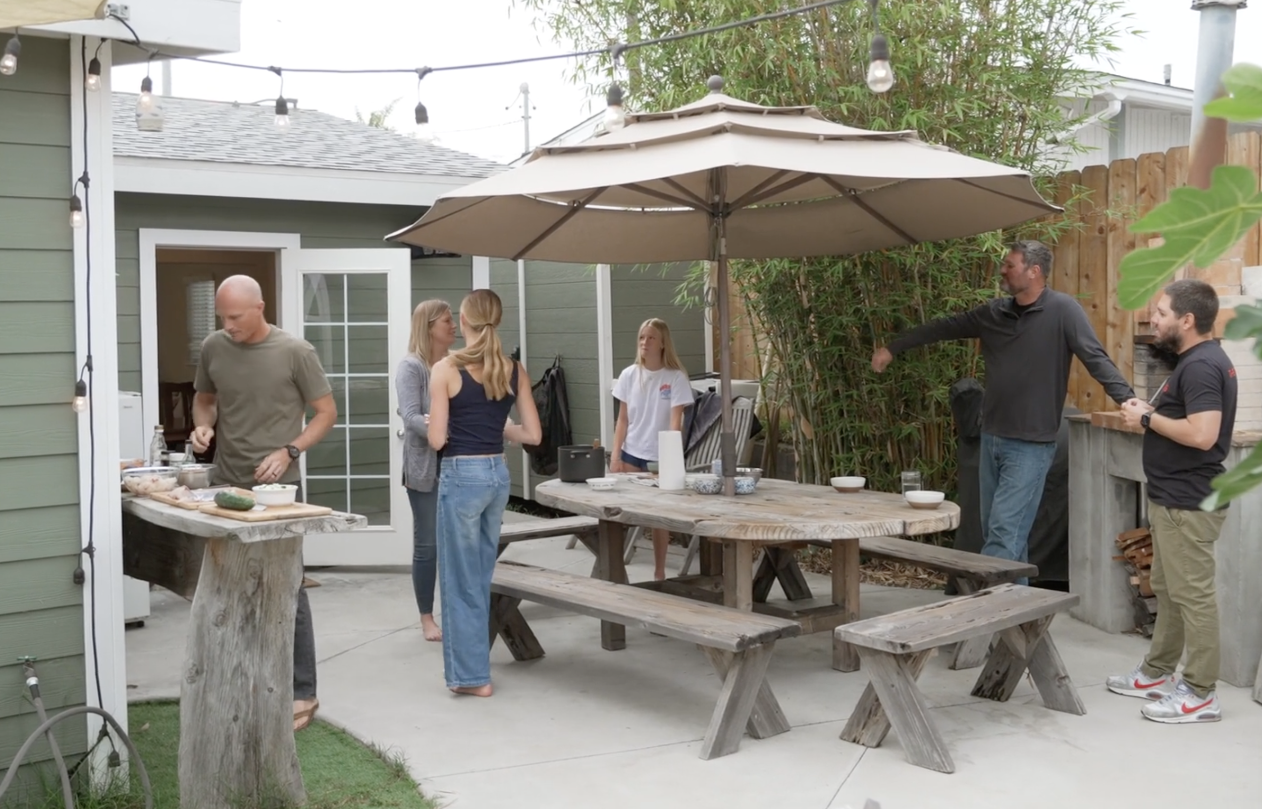 People gathered in a backyard patio with a large umbrella, preparing and socializing for a barbecue.