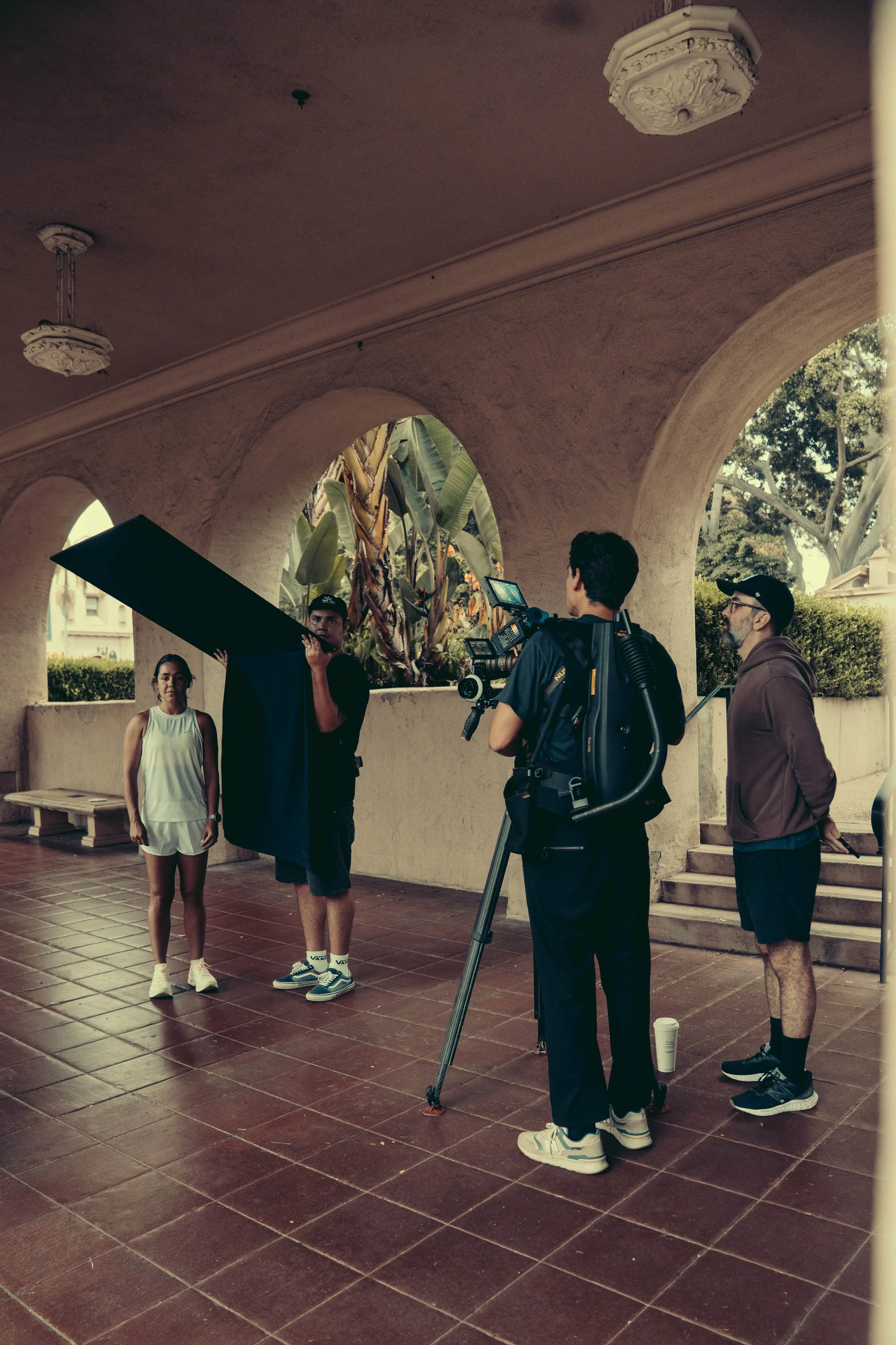 A film crew filming a scene with a female actor in an outdoor location with stucco walls and arches. Crew members are operating equipment, and one person is holding a reflector. The scene appears to be a casual interview or shoot in a warm, sunny env