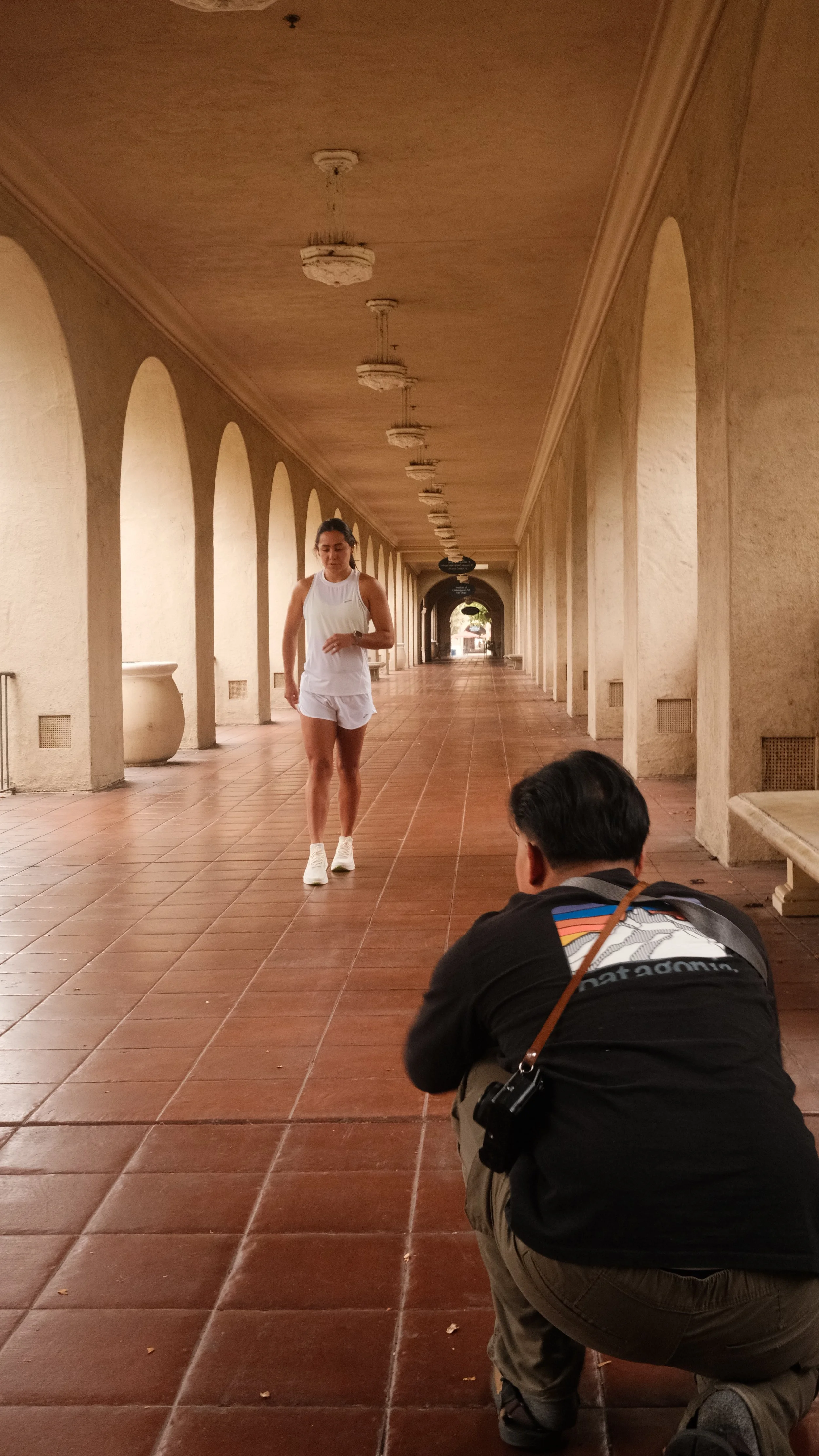 A woman in athletic clothing walking in a covered corridor with arches, while a man crouches taking a photo or video of her.
