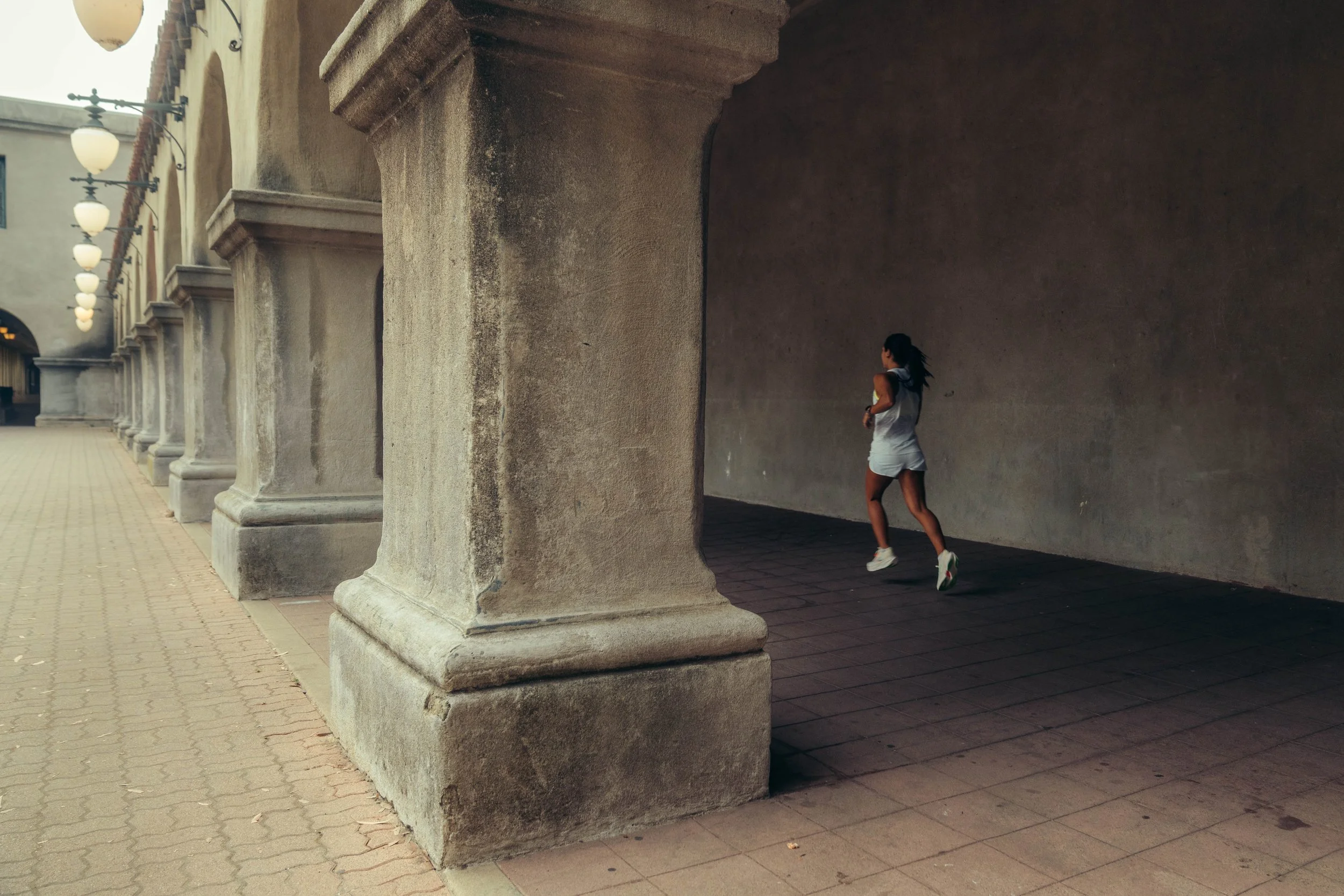 A person jogging through an outdoor corridor under a row of stone columns with arches. The corridor has hanging lamps and a brick roof, with a wall on the right and a paved walkway on the left.
