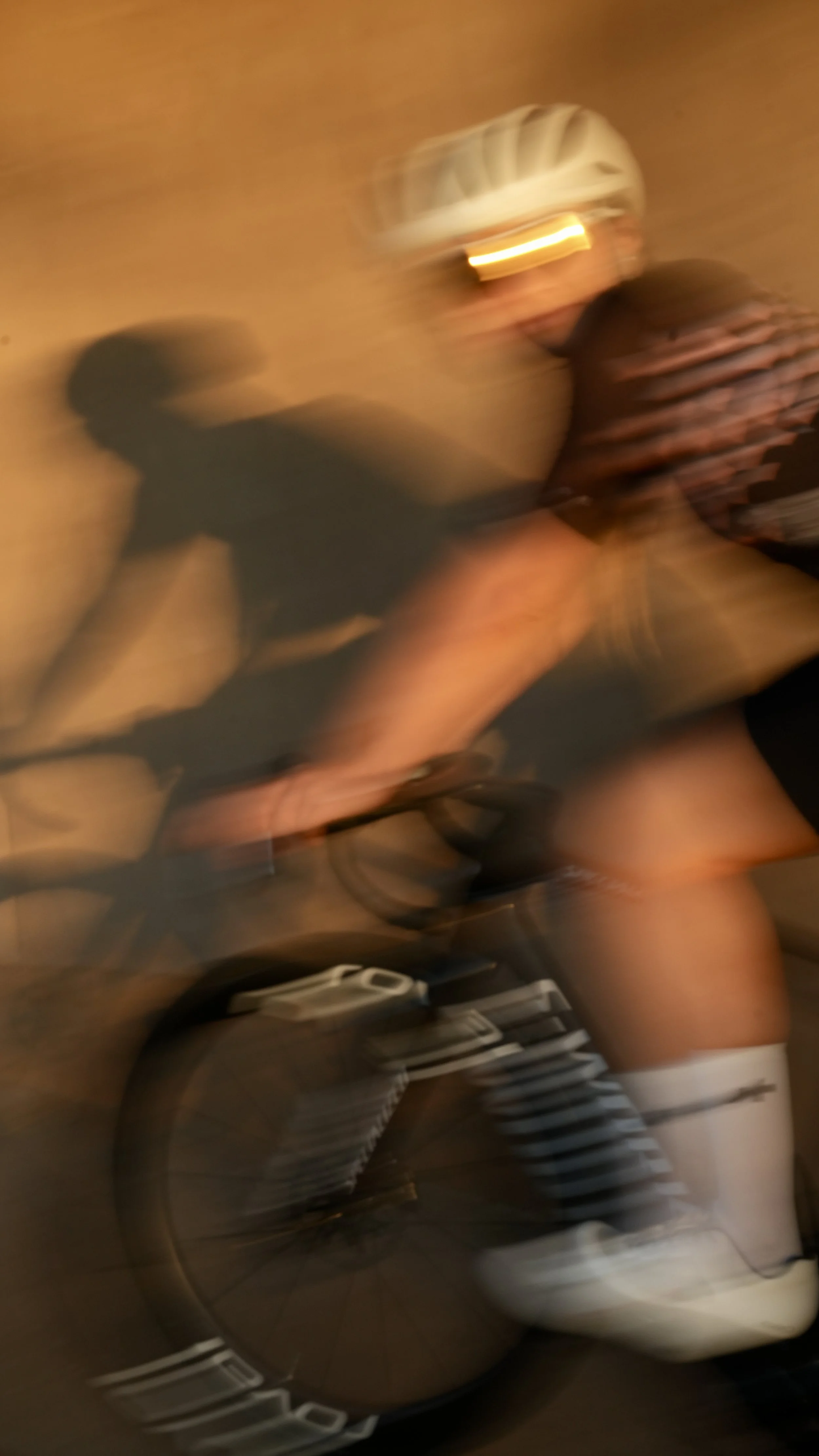 Blurred image of a person wearing a bicycle helmet, sunglasses, striped shirt, and shorts, riding a bicycle indoors.