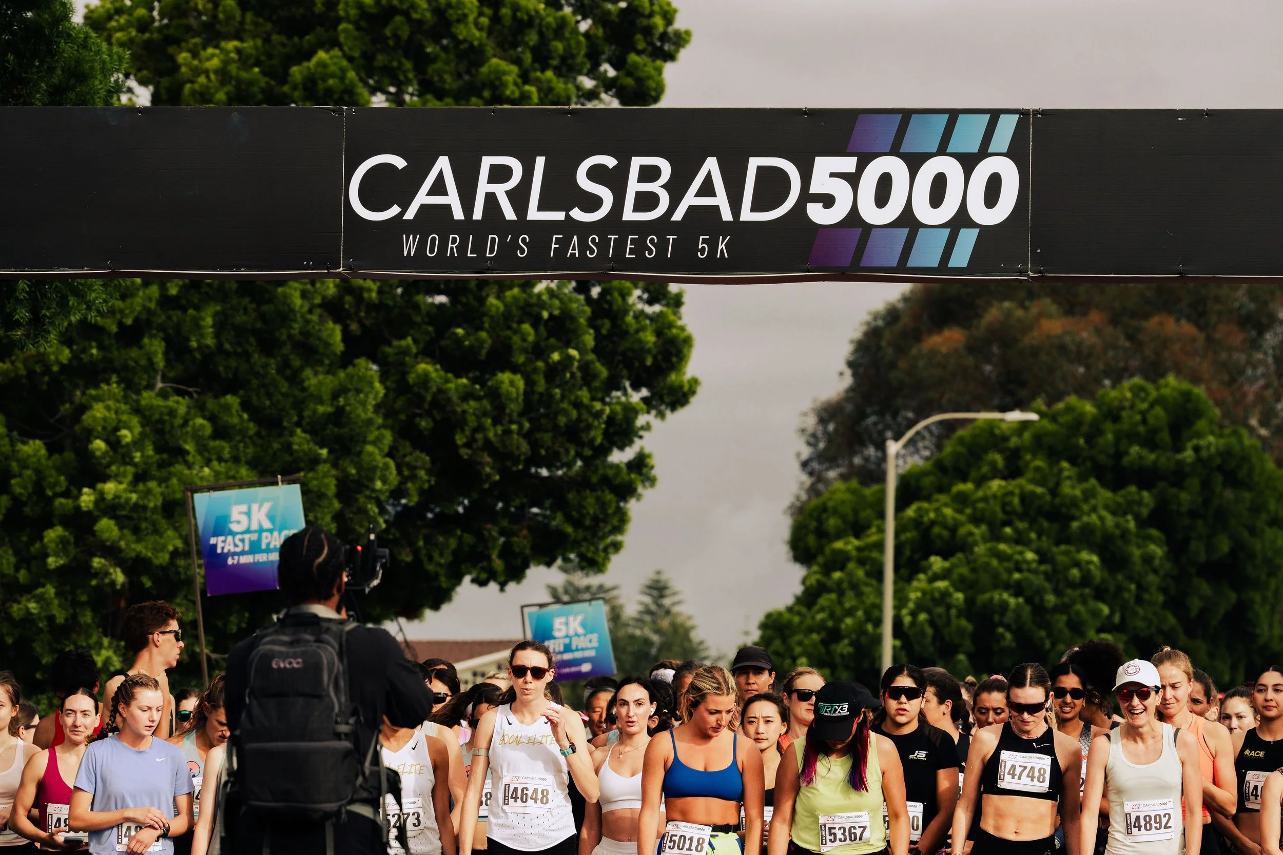 A large group of runners gathered at a race event, with a banner overhead reading 'CARLSBAD 5000, World's Fastest 5K.' People are wearing athletic clothes and race bibs, with trees visible in the background.