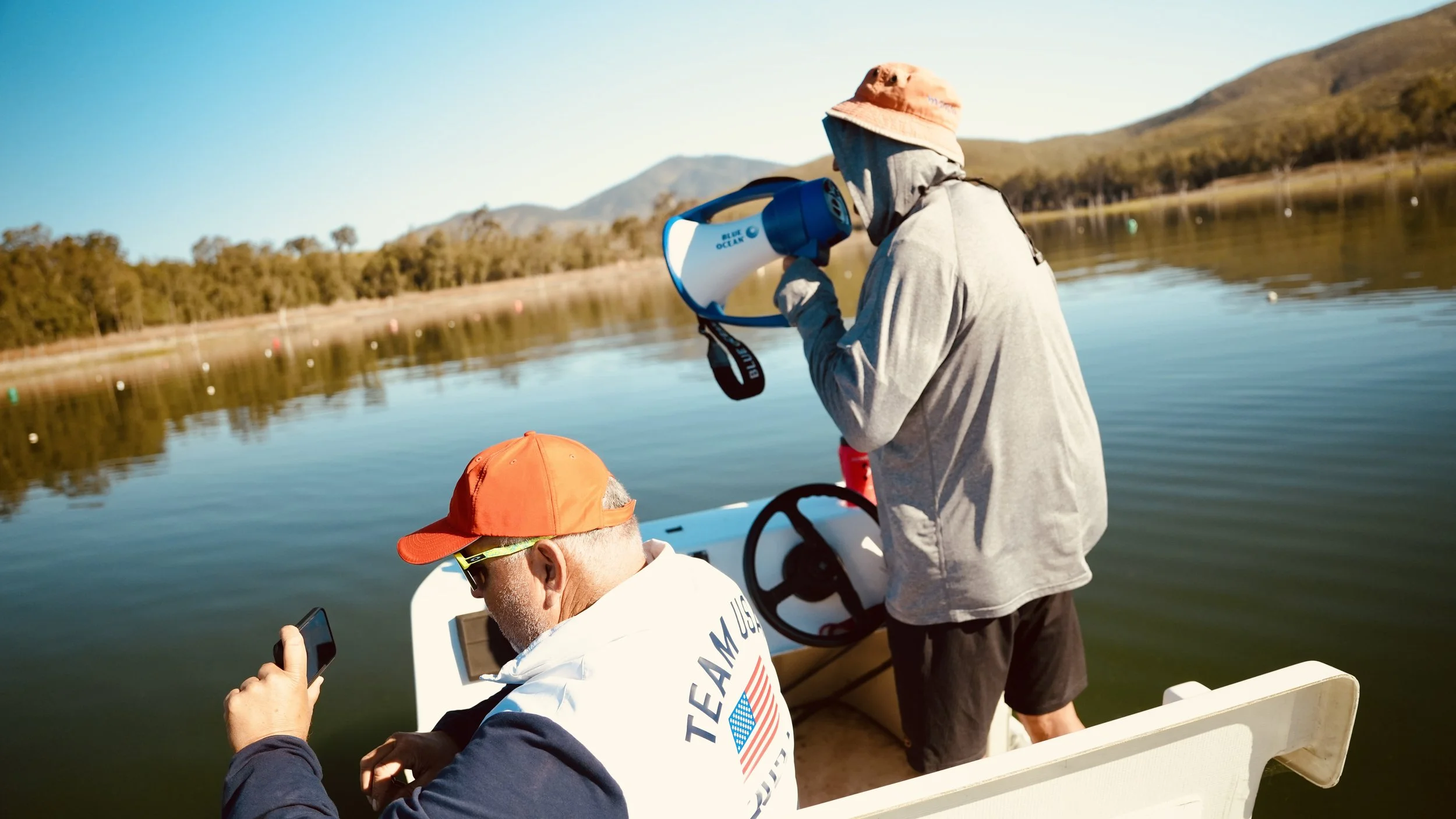 Two men on a boat on a lake, one wearing an orange cap and using a smartphone, the other holding a megaphone wearing a beige hat, gray hoodie, and black shorts, with mountains and trees in the background.