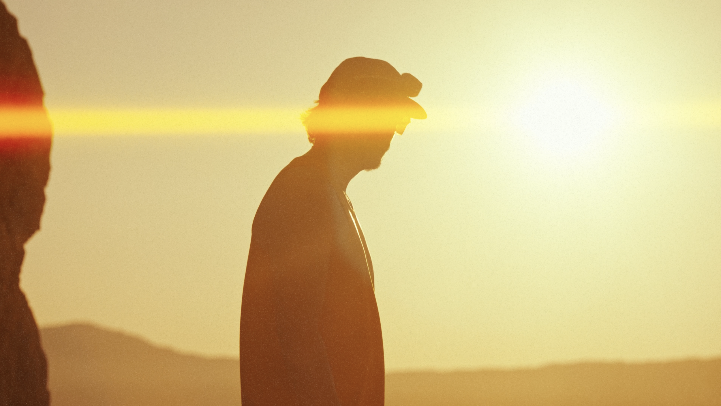 A person wearing a hat walking past a sunset with the sun casting a horizontal yellow-orange light across the scene.