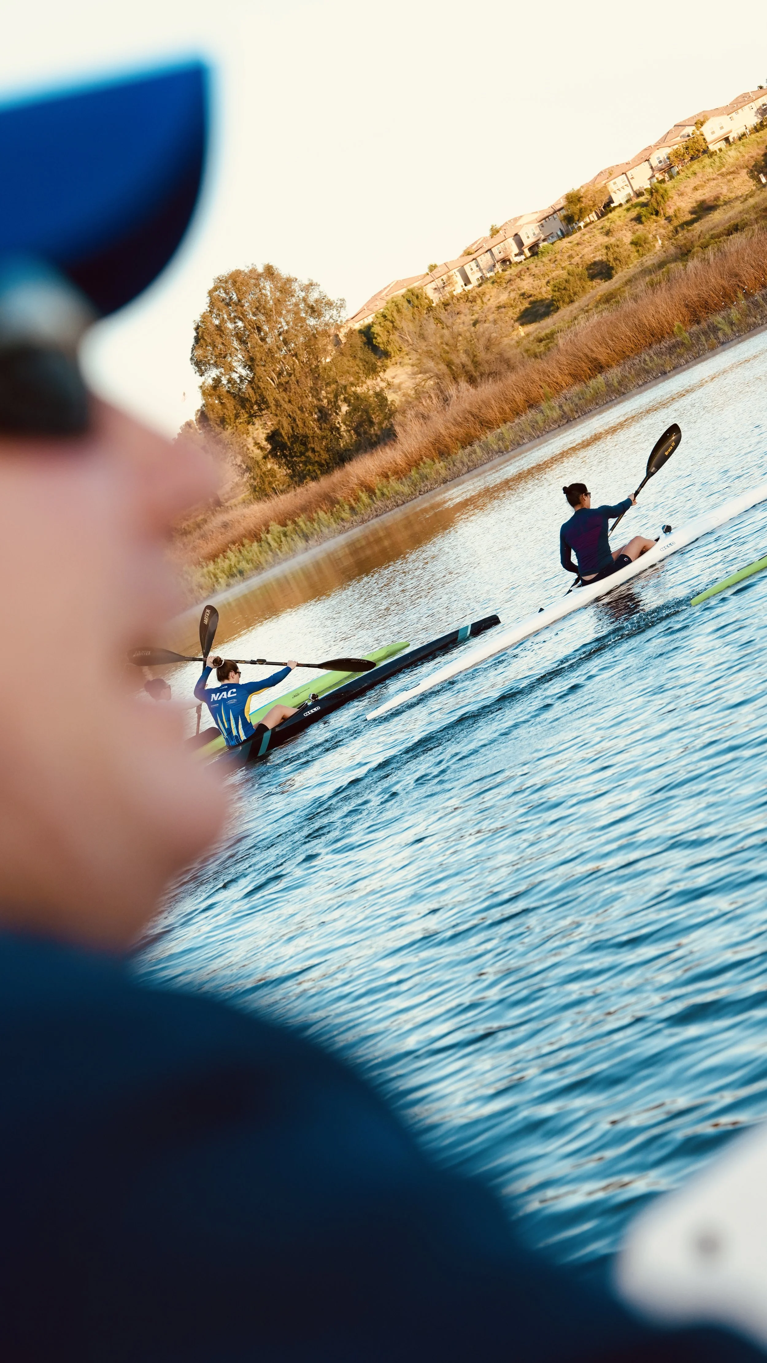 Two people paddleboarding on a calm body of water during sunset, with houses on a hill and trees in the background, partly obscured by a person's finger in the foreground.