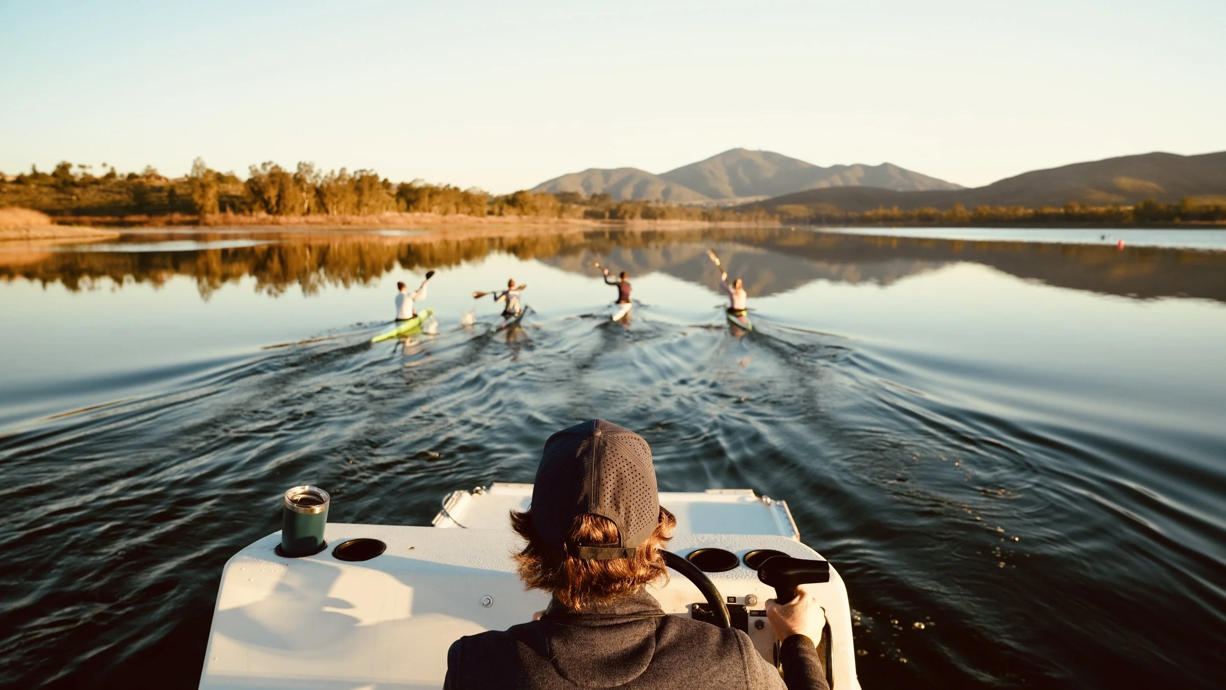Person steering a boat on calm water, with four people kayaking in the background, trees, mountains, and clear sky reflection on the water.