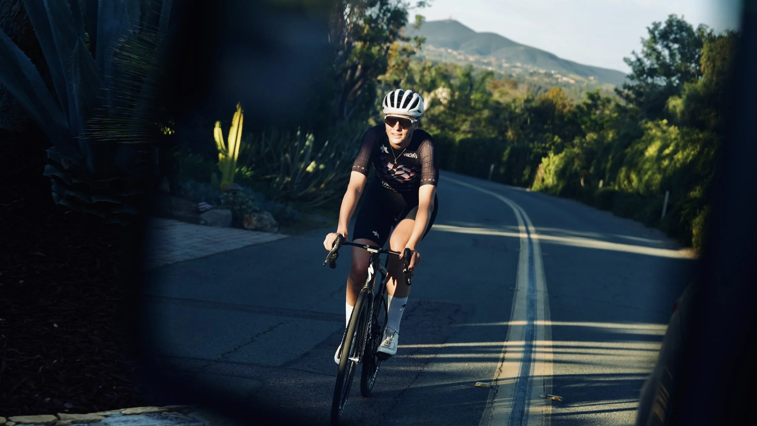 A woman riding a bicycle on a mountain road wearing a helmet, sunglasses, and athletic clothing with green trees and mountains in the background.